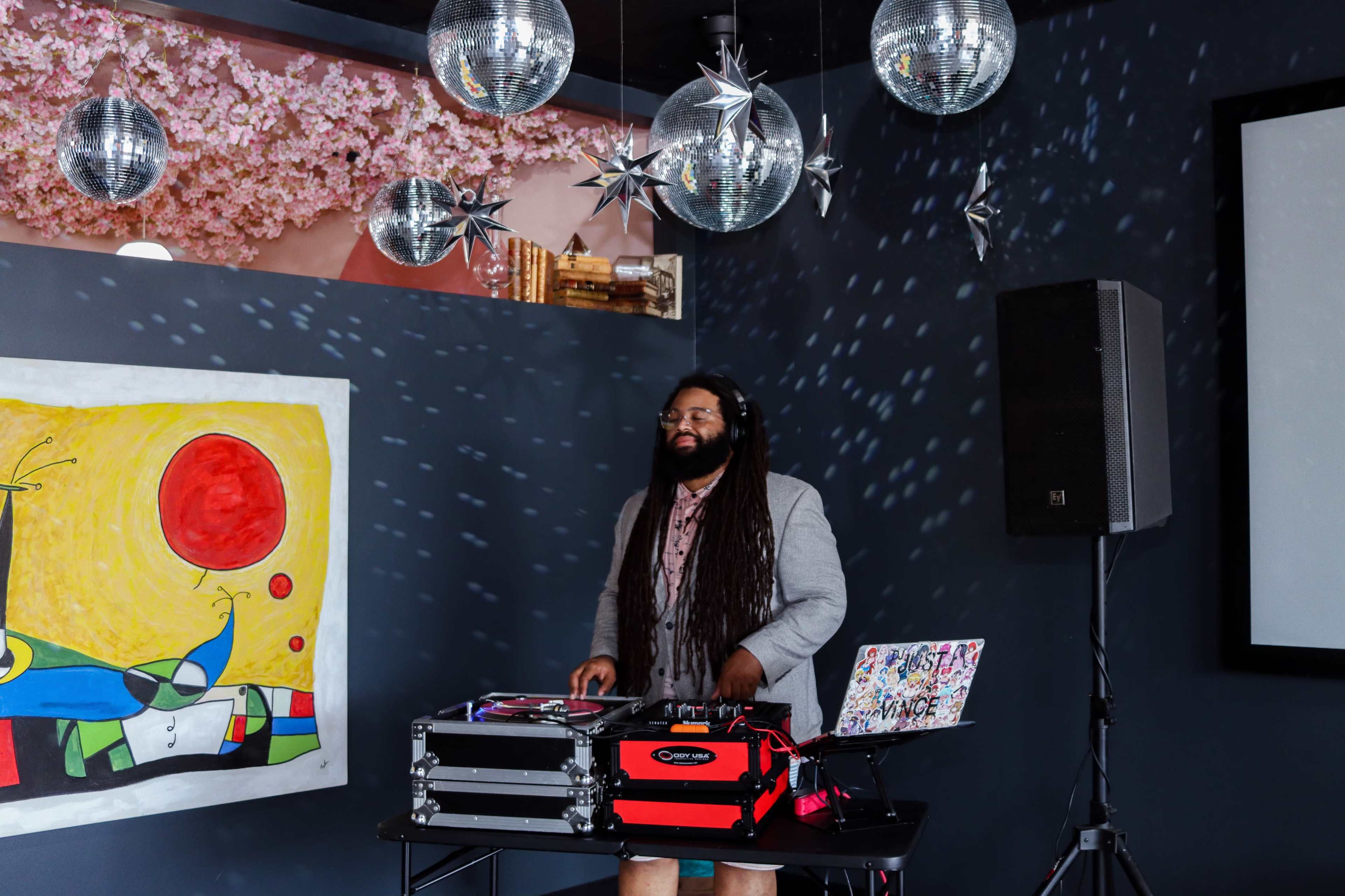 A DJ mixes music at a venue adorned with disco balls and decorative lights, while a vibrant artwork hangs on the wall.