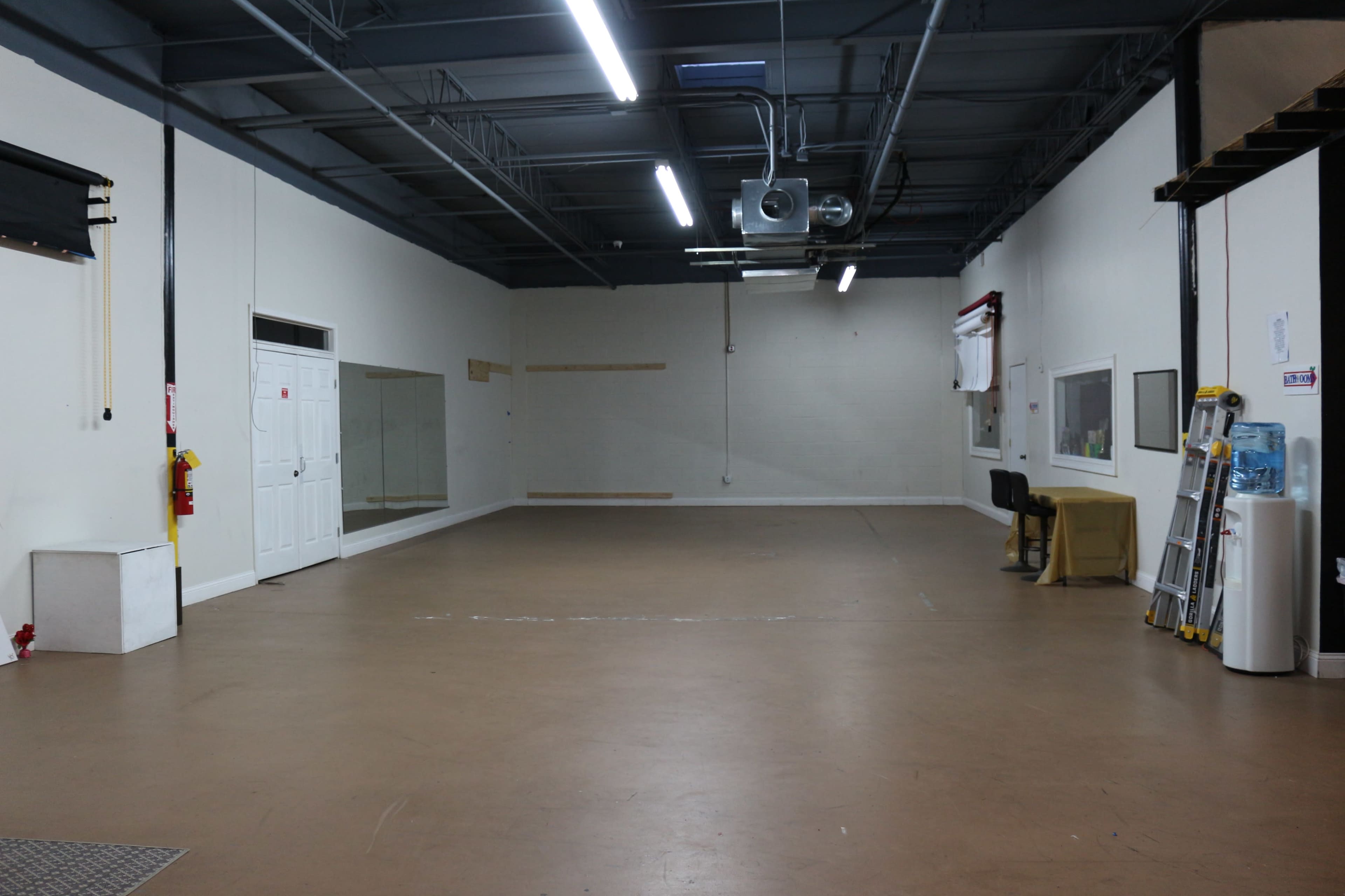 A large, empty room with beige floors, white walls, overhead lights, and a few simple items like a chair and a ladder.
