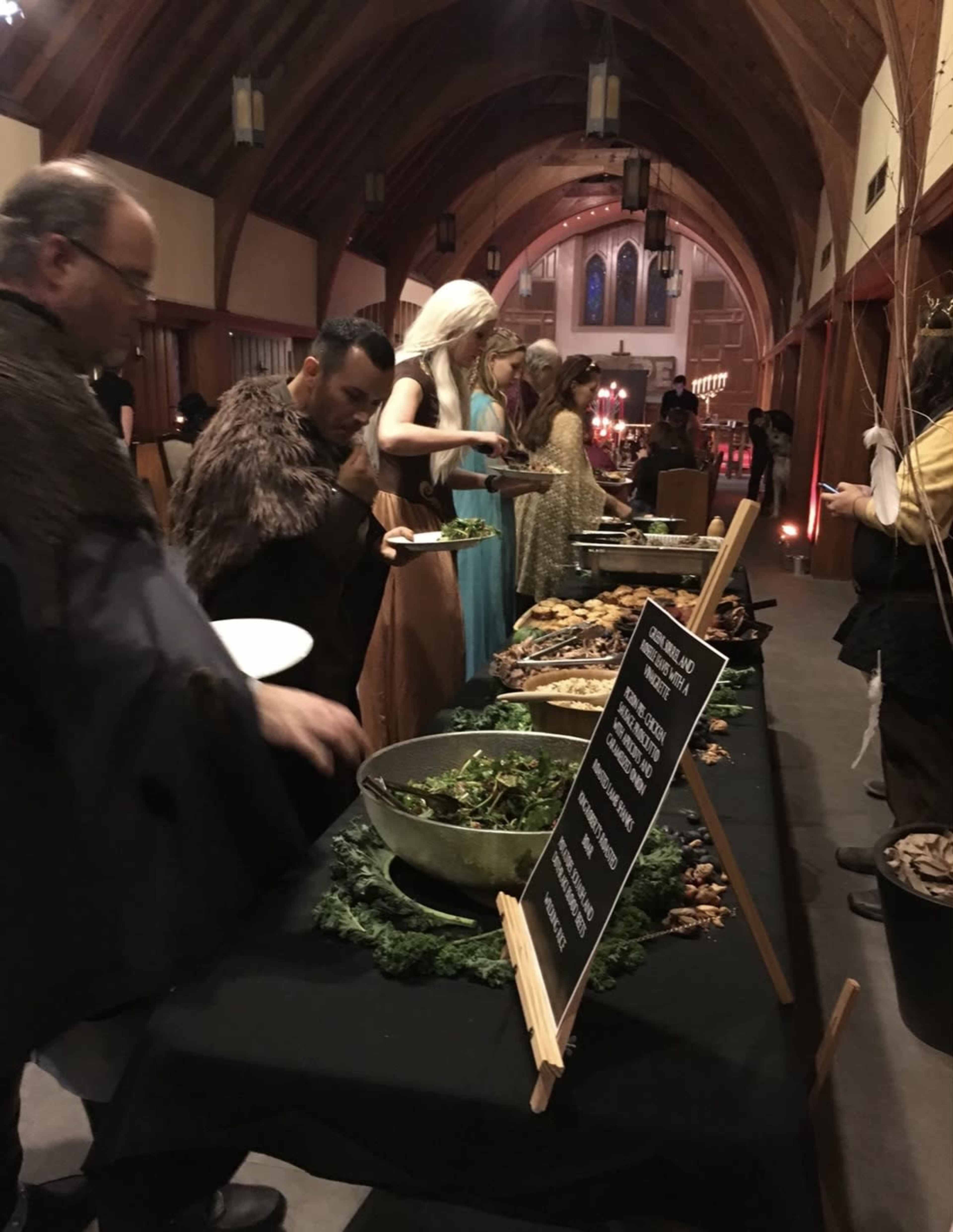 A group of individuals dressed in costumes serves themselves from a long buffet table filled with food in a dimly lit hall with wooden arches.
