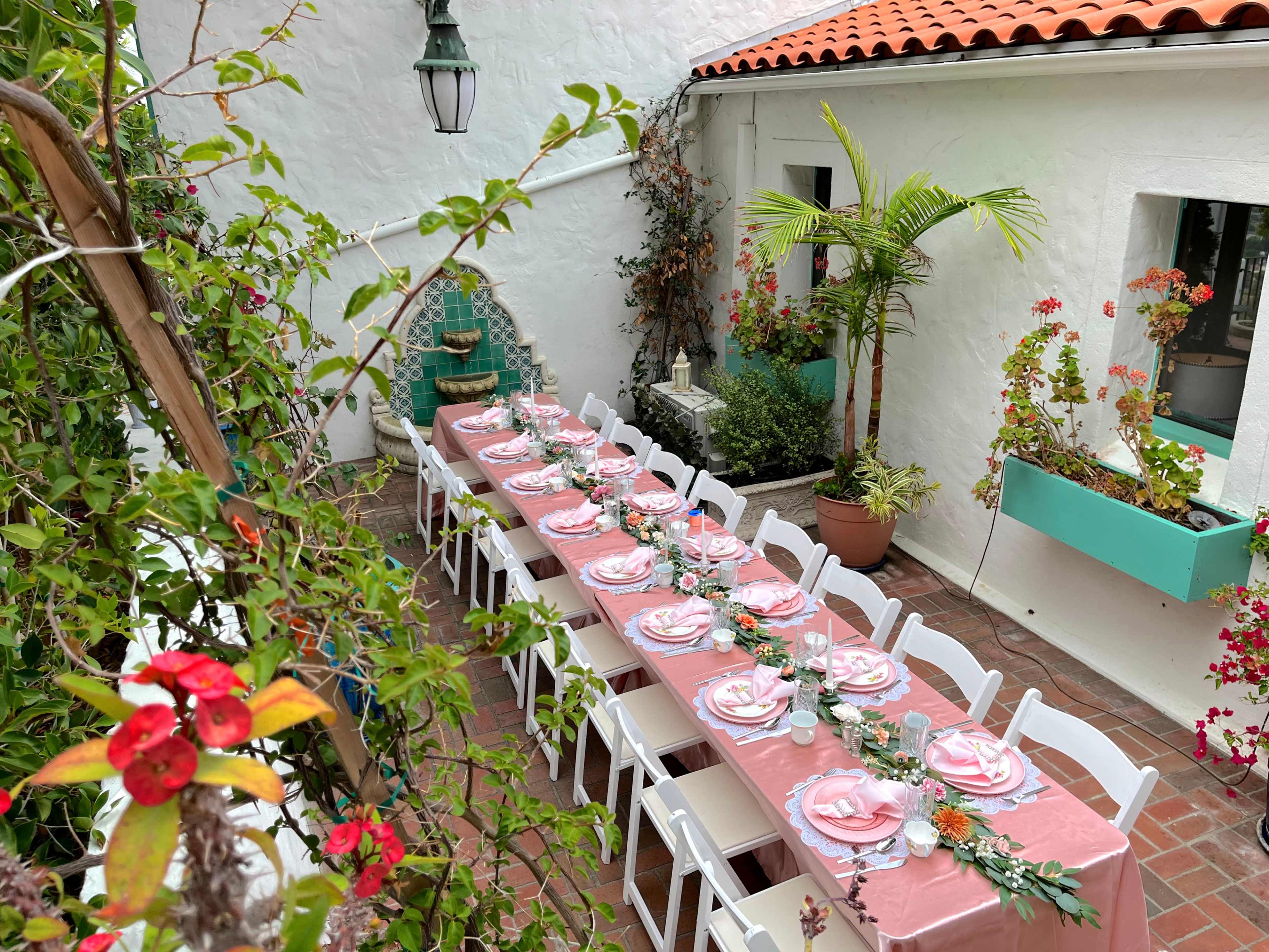 A long table is set for a gathering in a courtyard, adorned with pink tablecloths and floral decorations, surrounded by potted plants and a white wall.