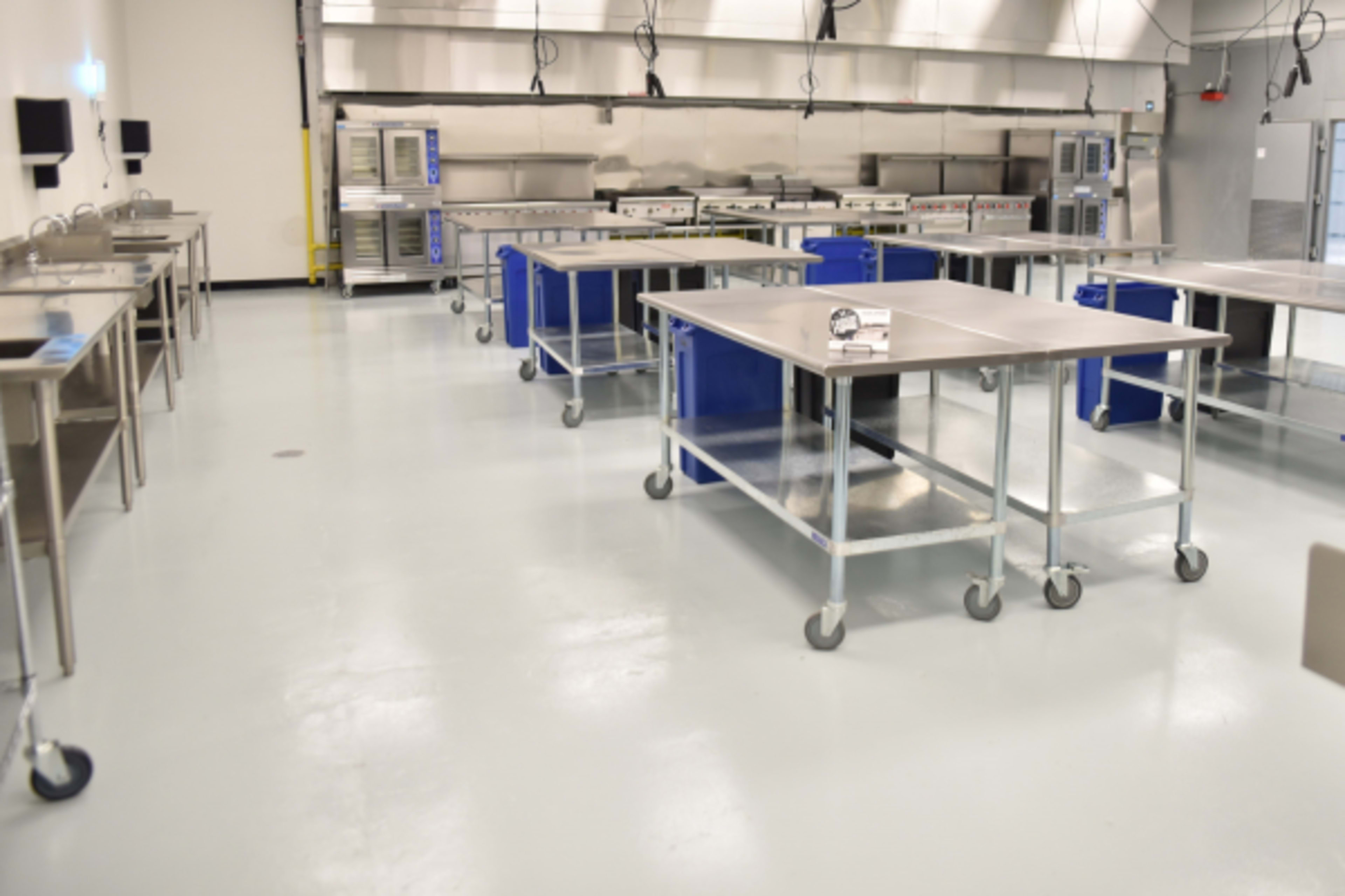 A spacious, industrial kitchen with stainless steel tables on wheels and a series of ovens in the background.