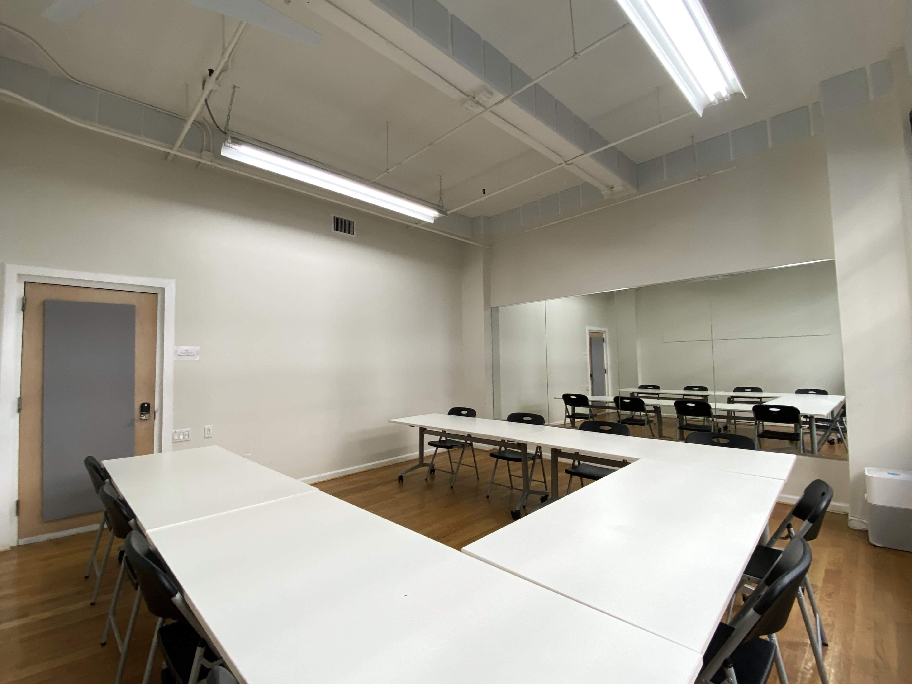 A meeting room with white tables arranged in a U-shape and black chairs positioned around them, with a large mirror on one wall.