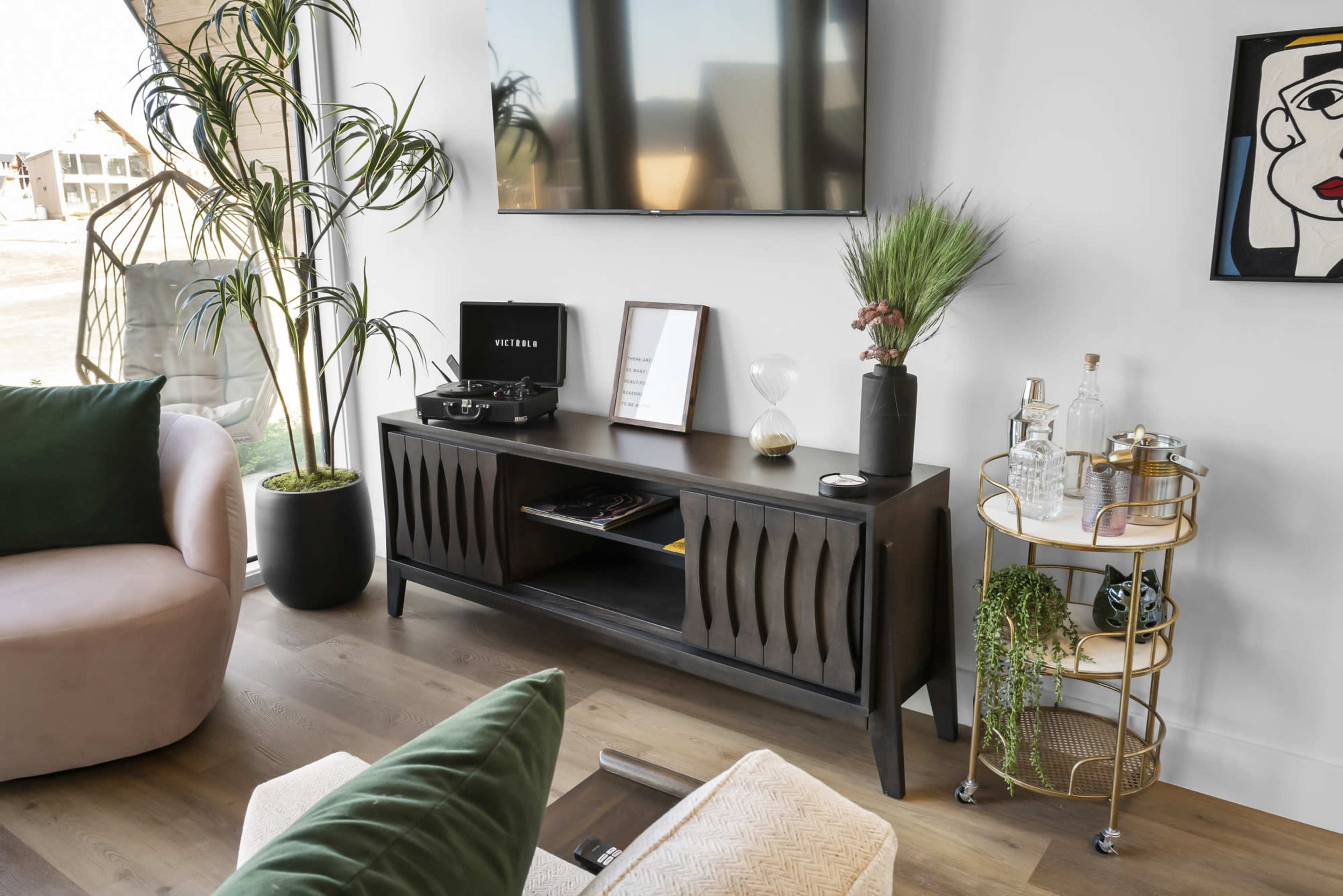A modern living room features a black media console under a mounted TV, surrounded by plants and decorative items.