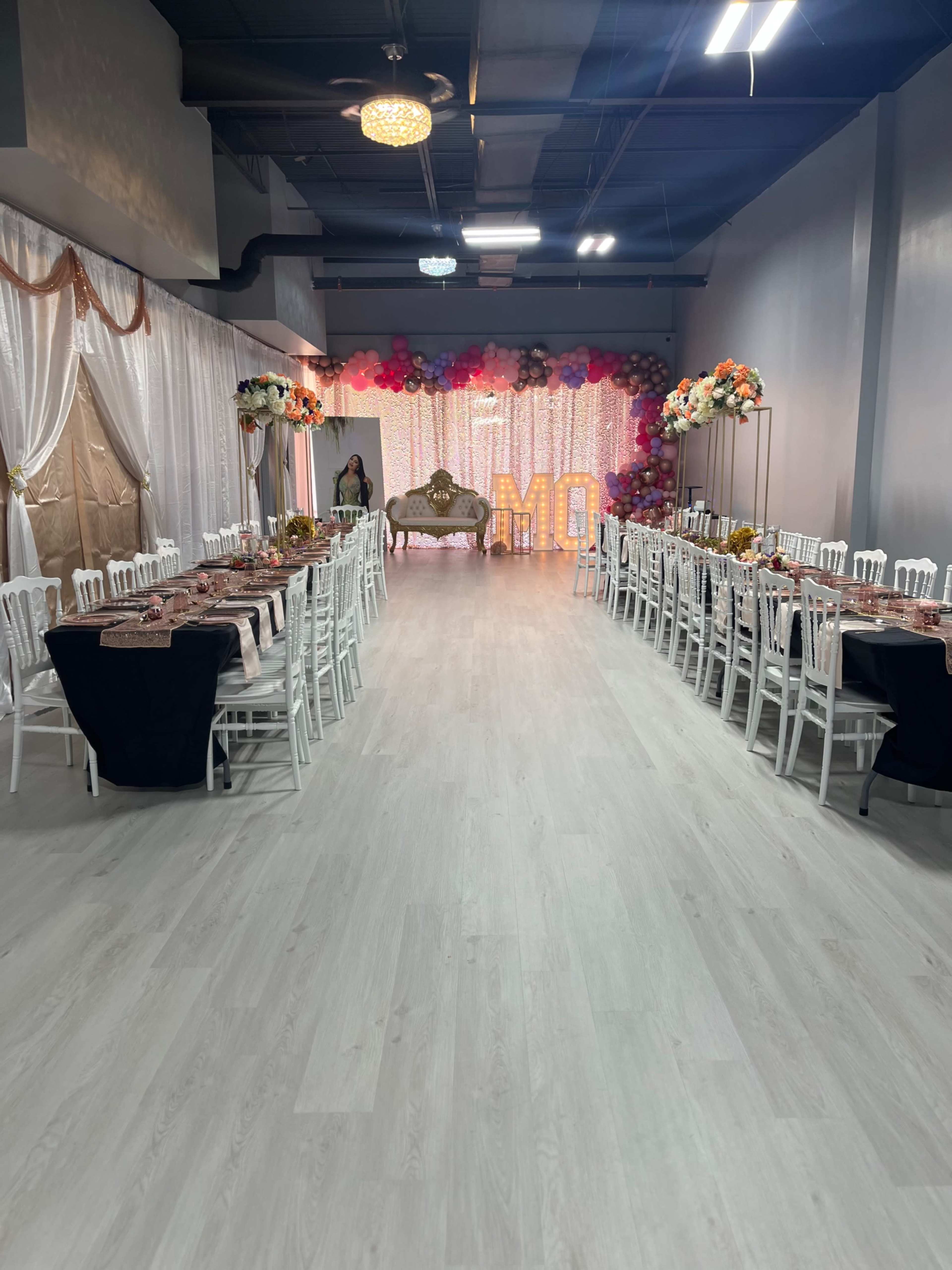 A spacious event venue set up with long tables and white chairs, decorated with floral arrangements and a sparkling backdrop.