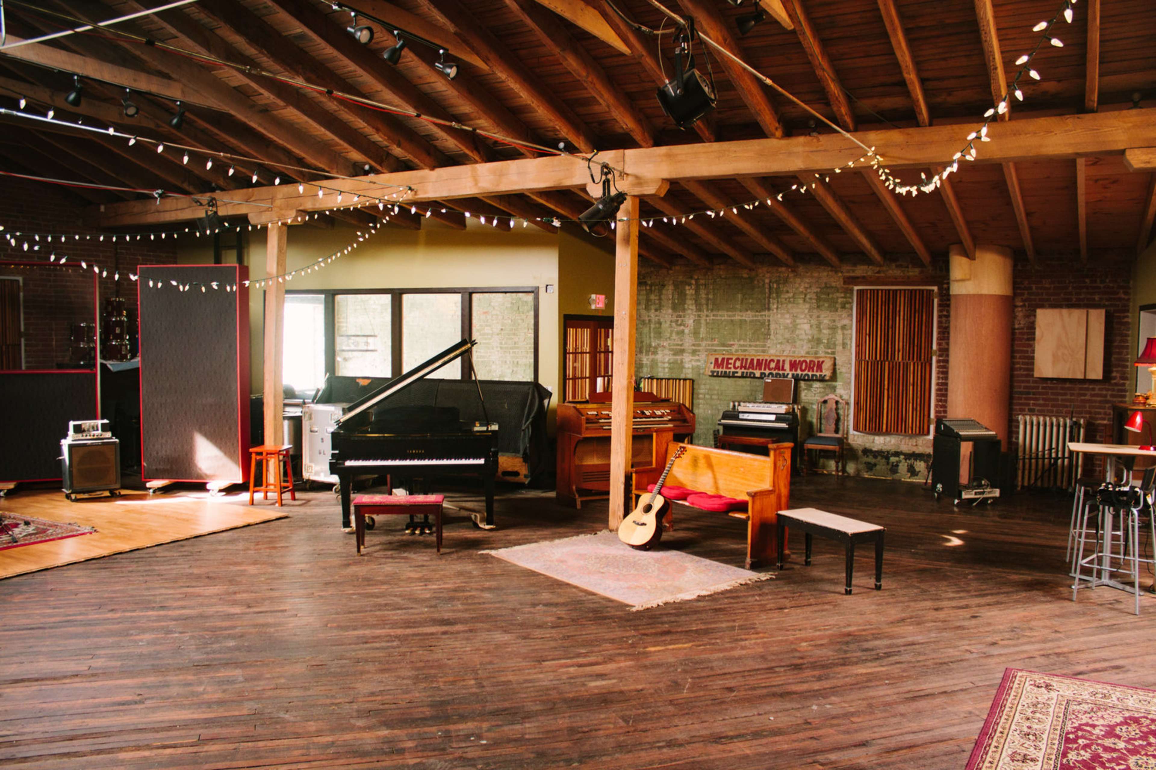 A spacious music studio featuring a grand piano, several amplifiers, a guitar resting on a wooden bench, and warm string lights overhead.
