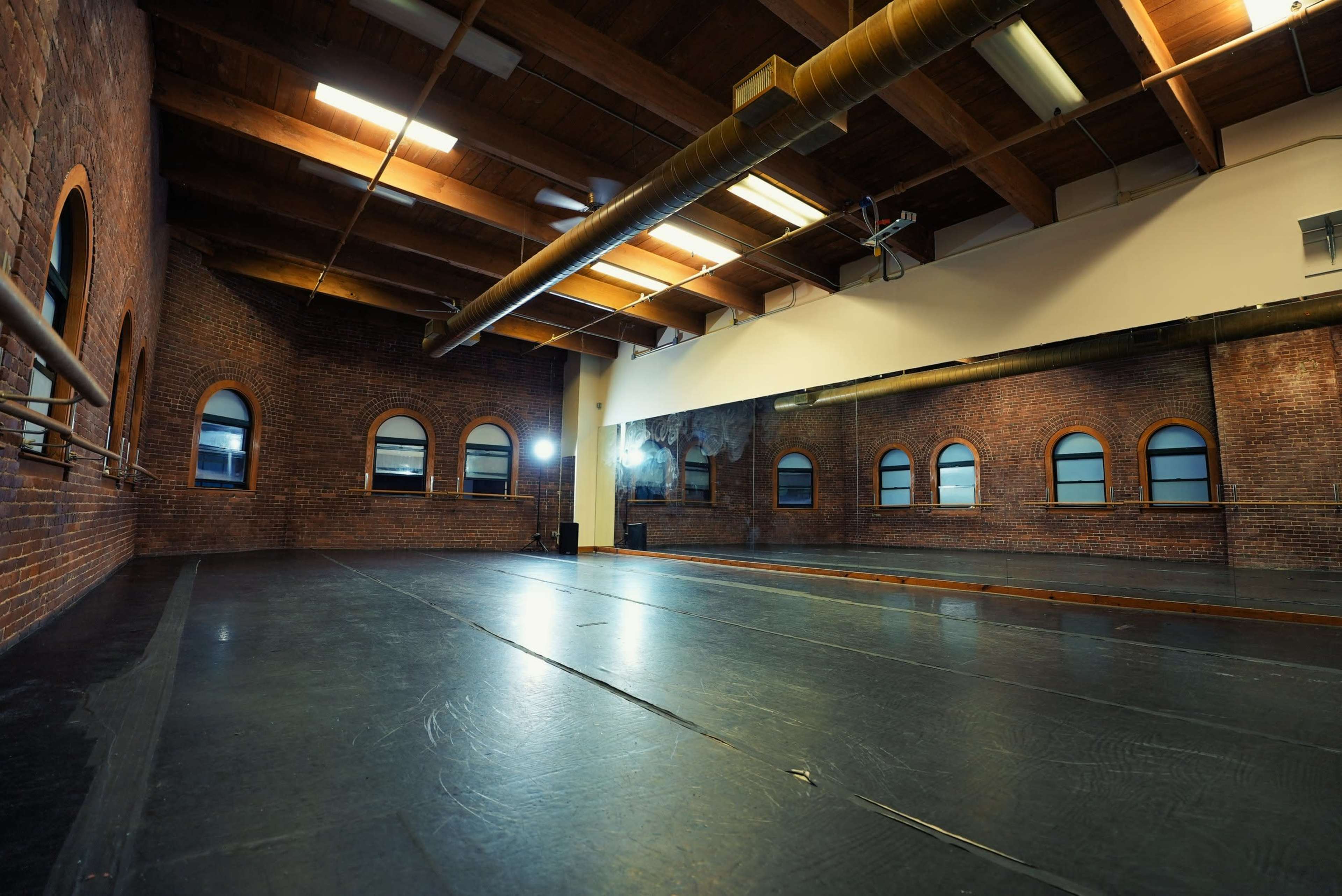 A spacious dance studio with brick walls, large windows, and a wooden ceiling.