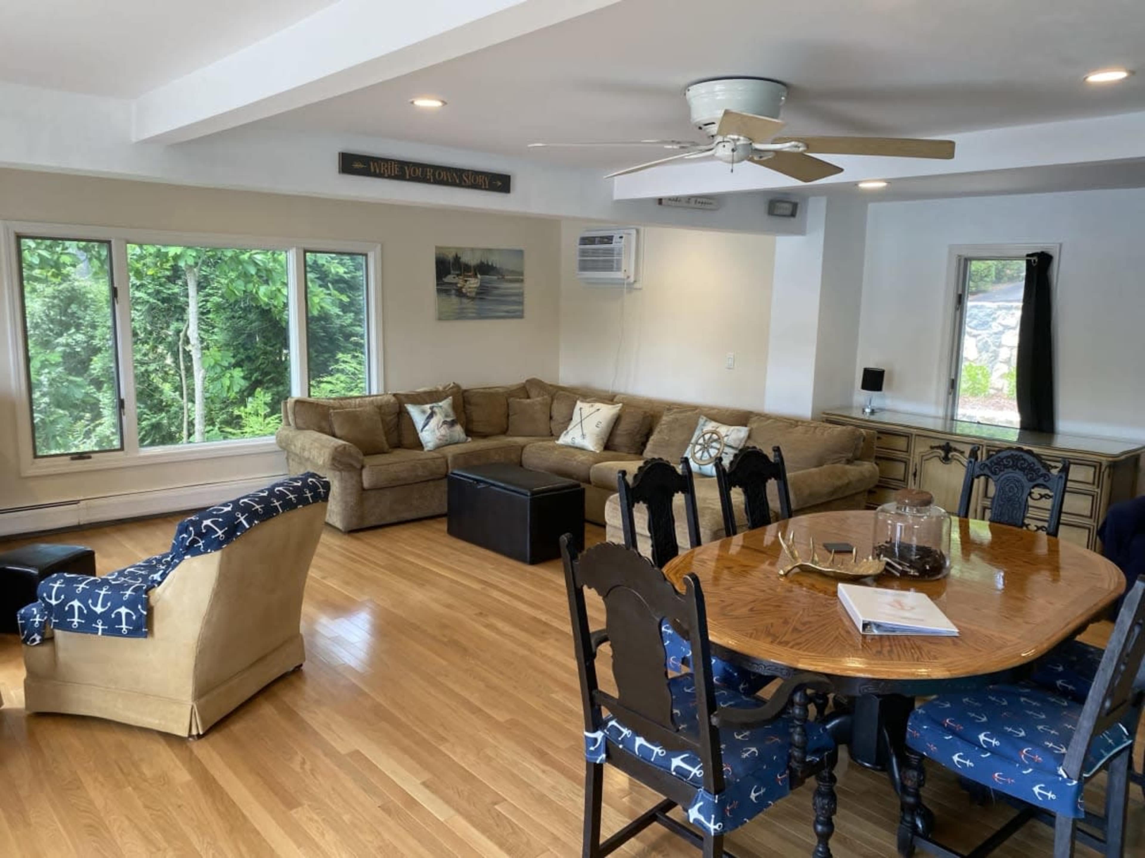 A living and dining area with a sofa, chairs, a coffee table, and a dining table surrounded by chairs, with large windows providing views of greenery outside.