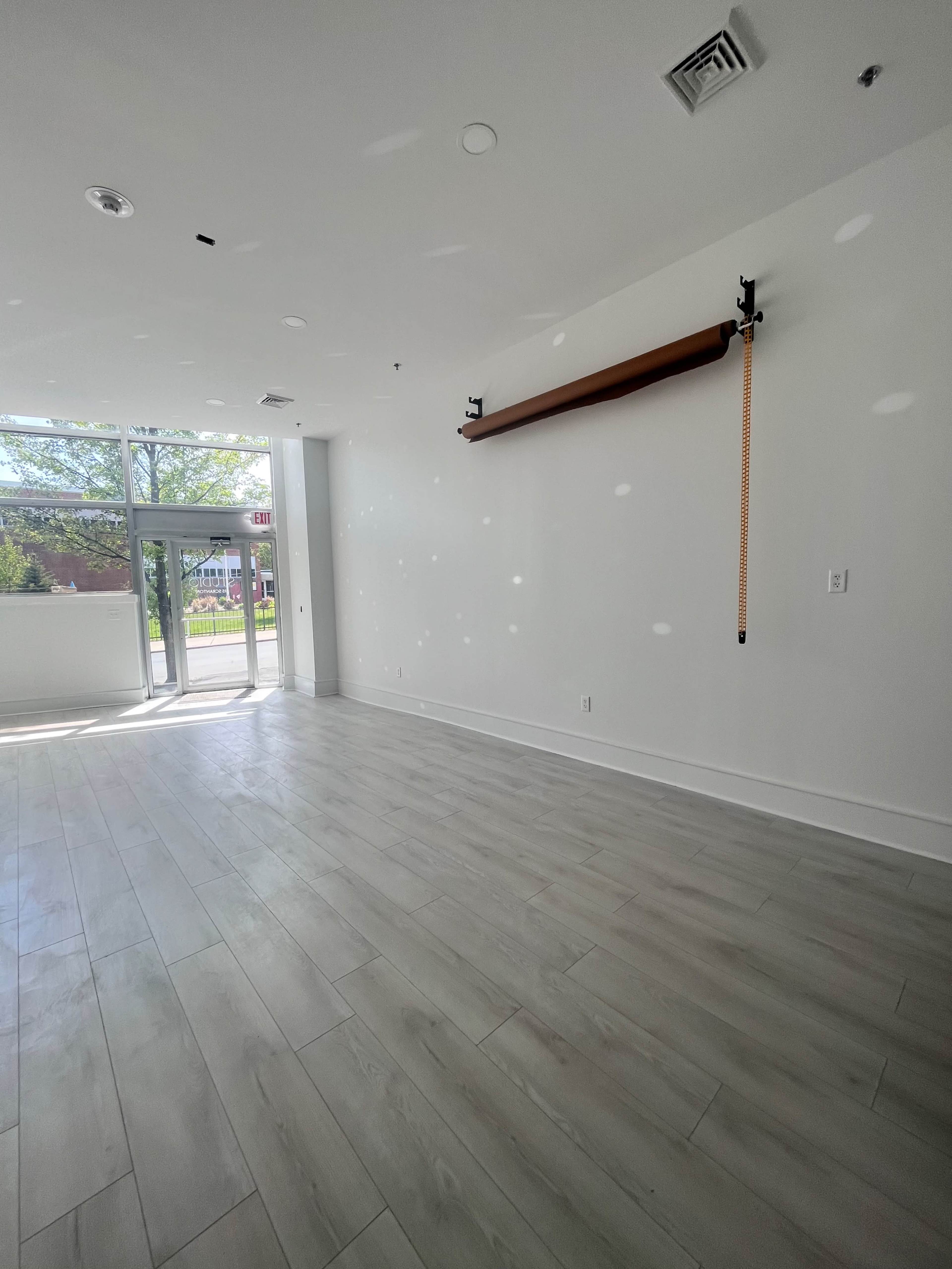 A vacant room with large windows letting in natural light, featuring light-colored tiled flooring and a wooden shelf mounted on the wall.