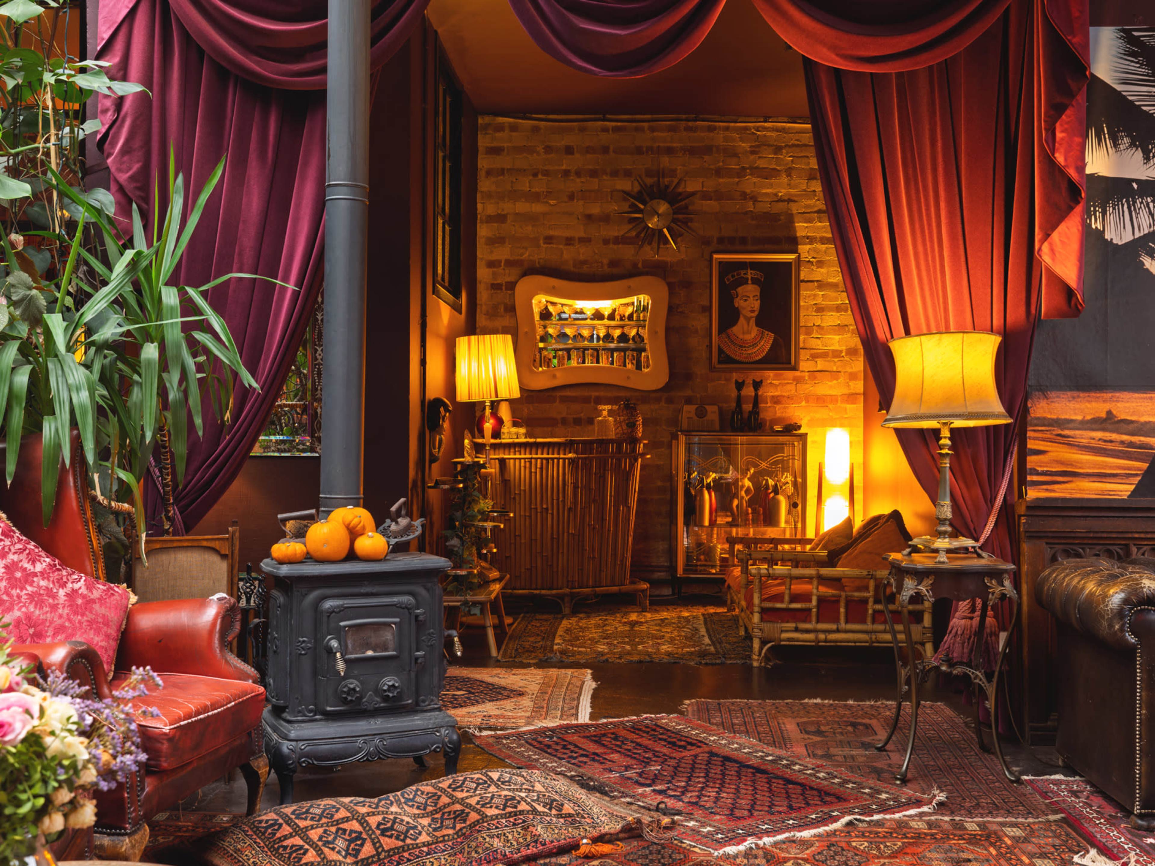 The image depicts a cozy, dimly lit room adorned with rich red drapes, a vintage wood-burning stove, and eclectic furnishings, including a framed portrait and vibrant plants.