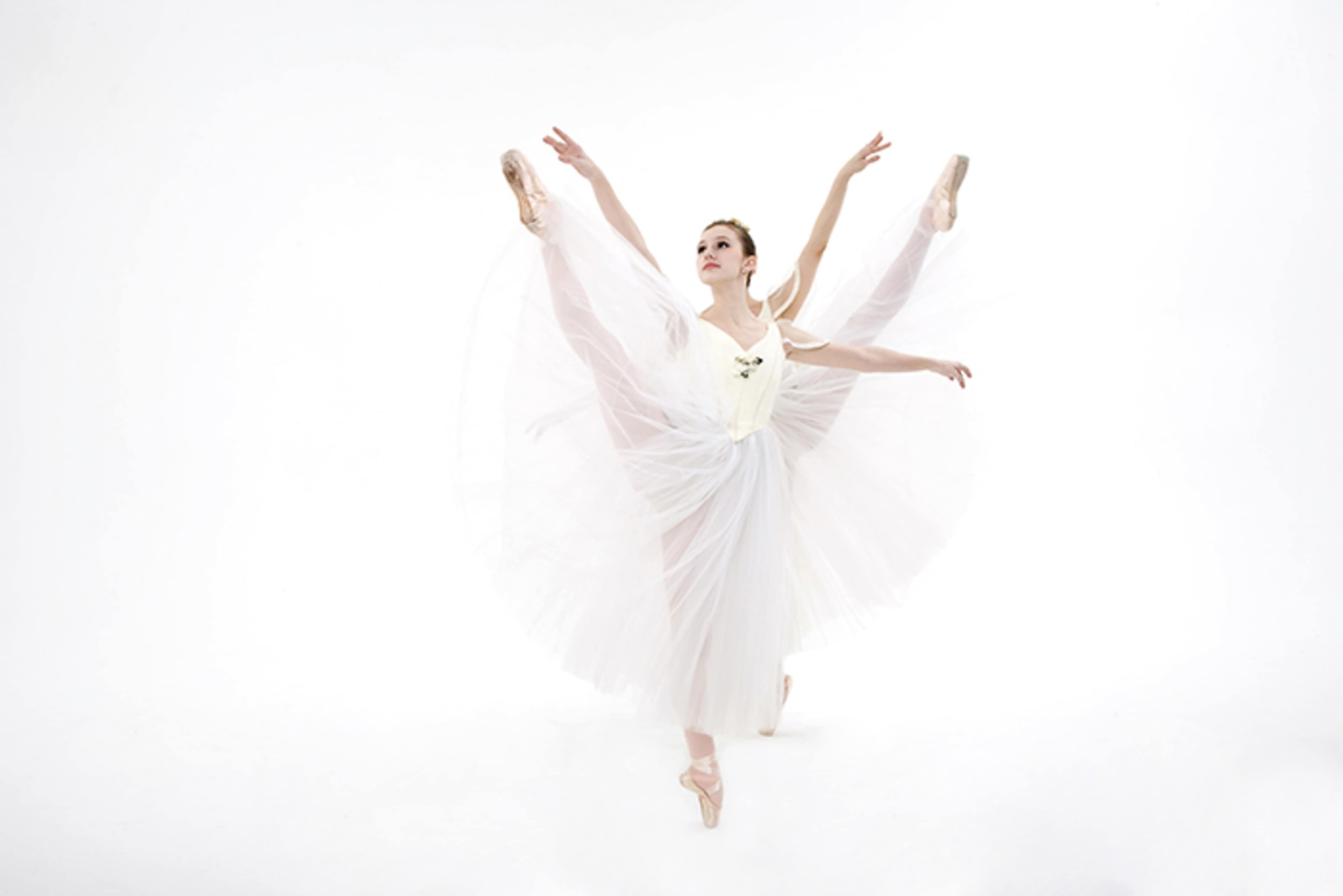 A ballet dancer strikes an elevated pose, showcasing her extended legs and flowing white tutu against a plain white background.