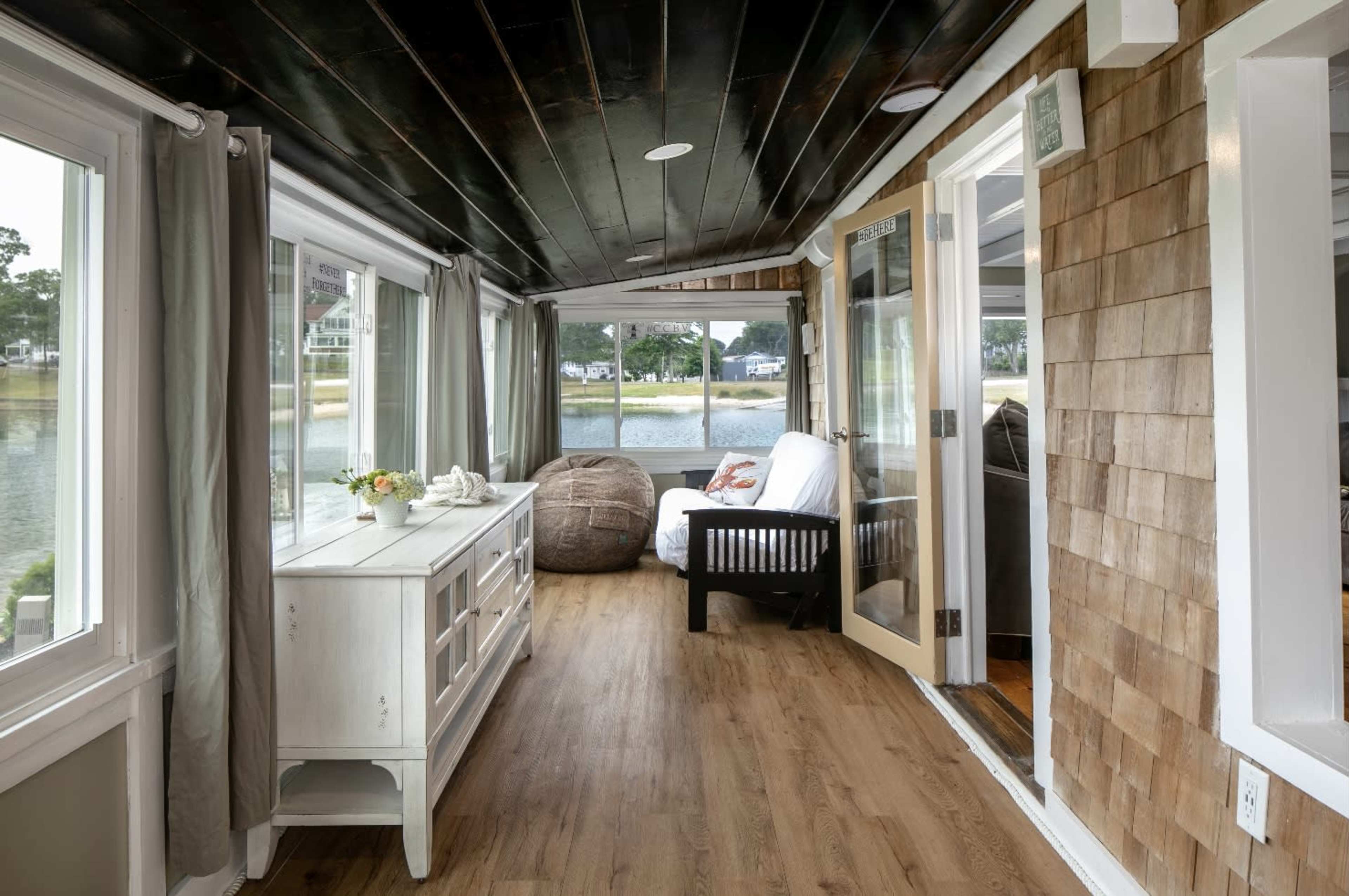 A sunroom with large windows overlooking a body of water, featuring wooden flooring, a white cabinet, a bean bag chair, and a black loveseat.