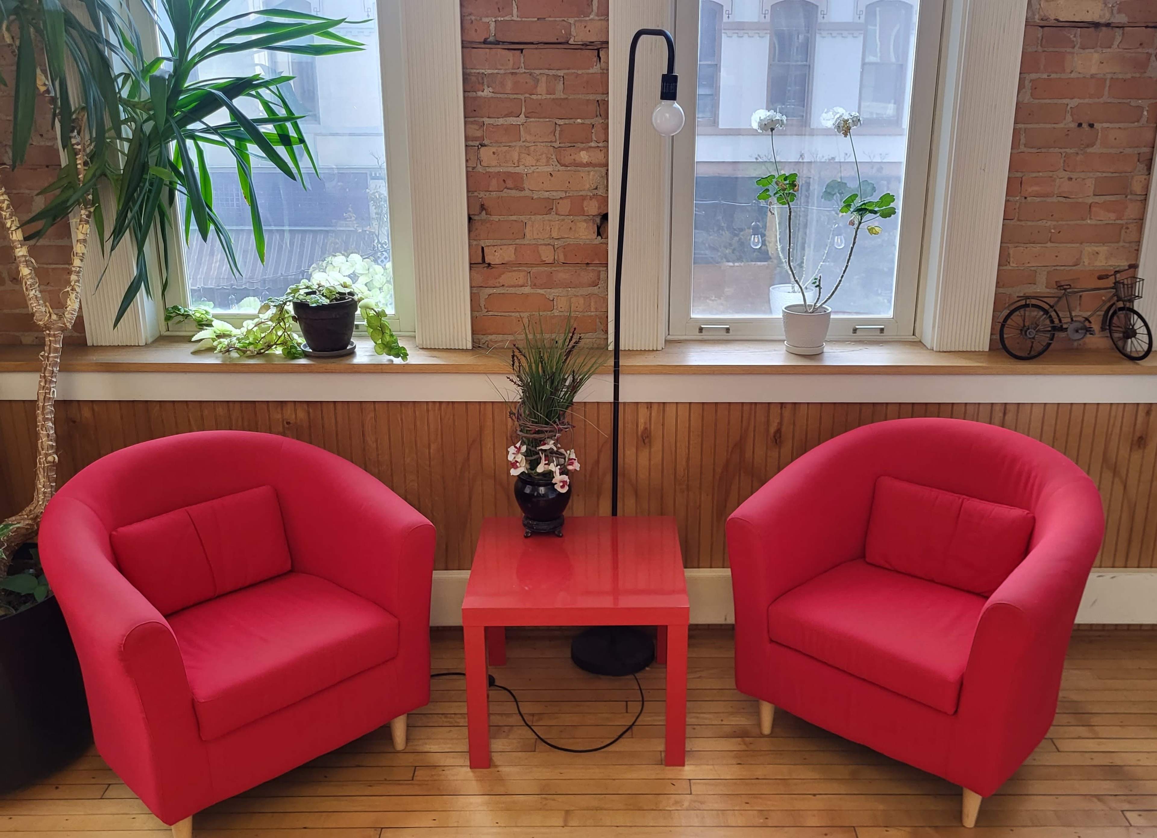 Two red armchairs flank a small red table beneath a lamp, with potted plants on the windowsill behind them.