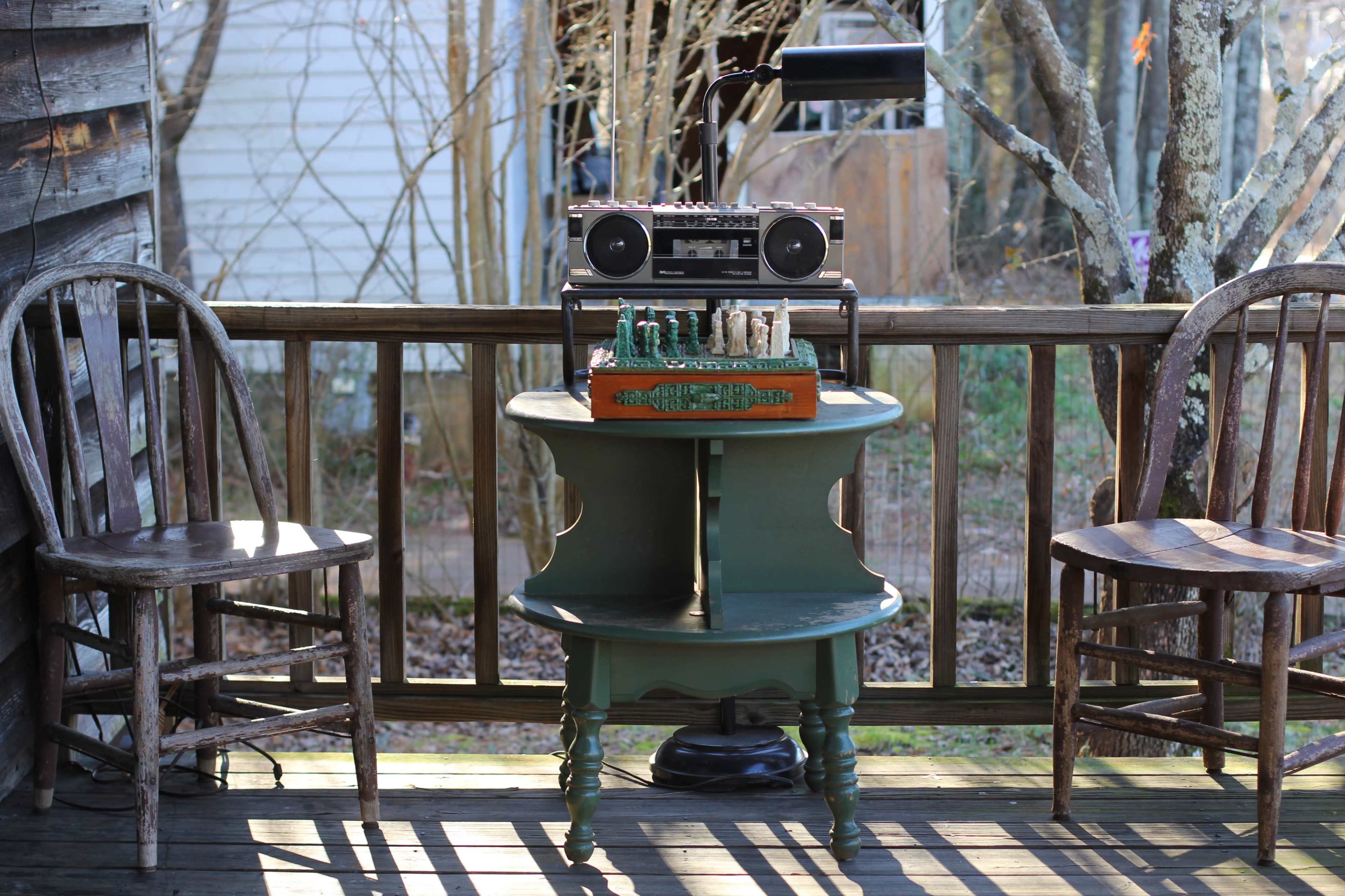 An old wooden porch features a round table with a vintage boombox and a decorative box, flanked by two mismatched chairs.