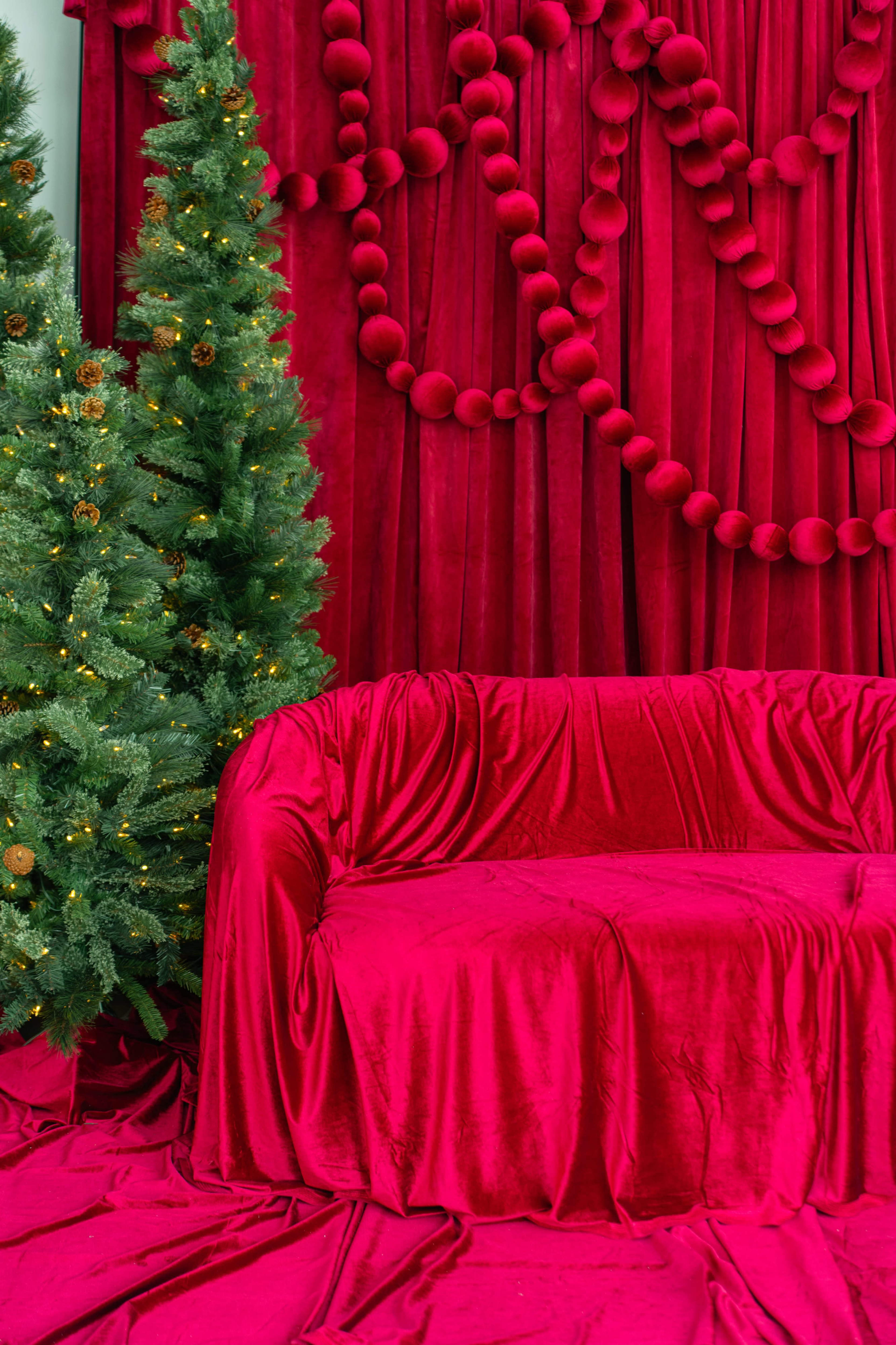 The image features a red-themed holiday setting with a plush red sofa draped in fabric, flanked by two Christmas trees decorated with lights.