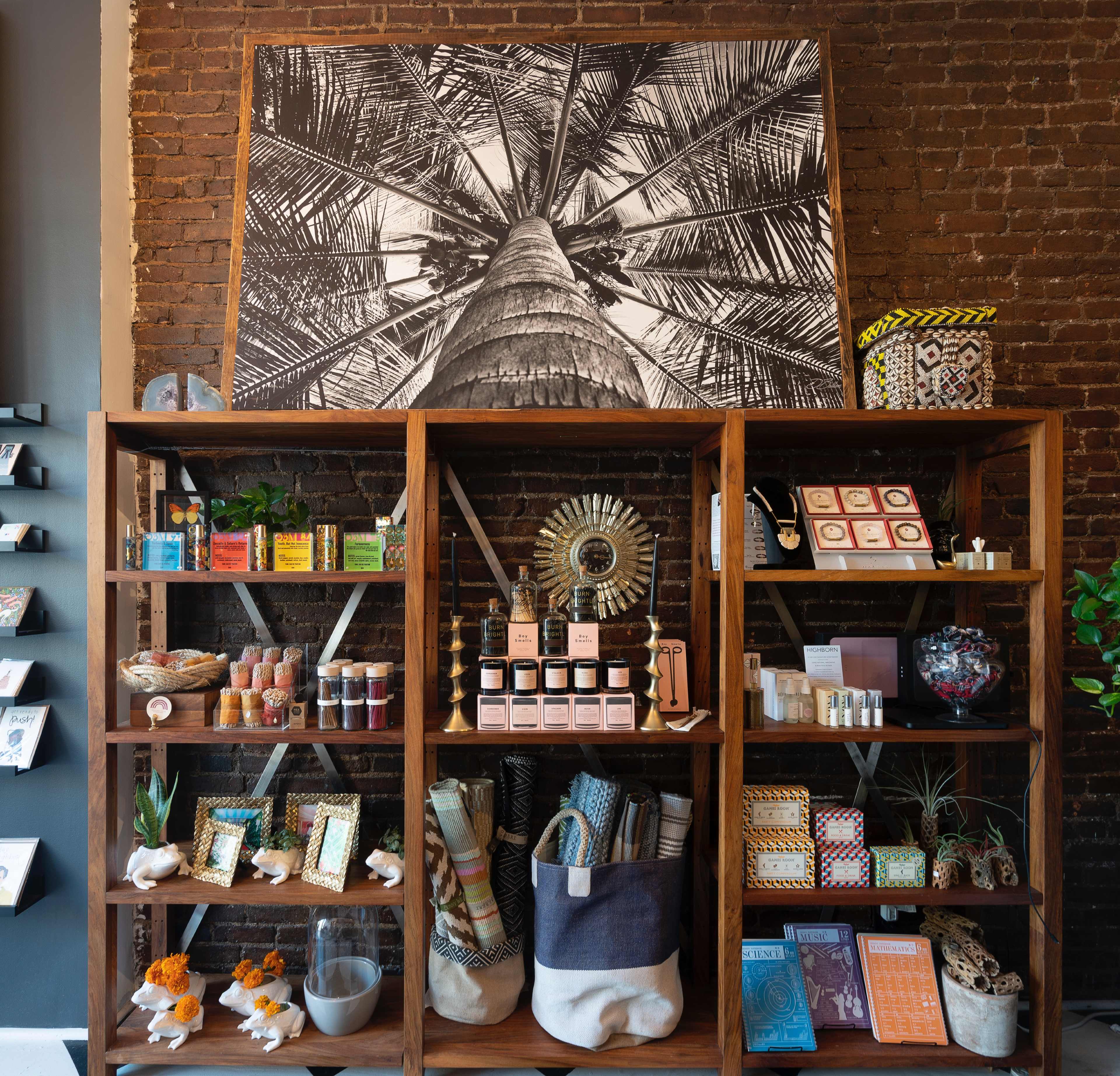A wooden shelf displaying a variety of colorful products, including candles, beauty items, and decorative accessories, against a backdrop of a large black-and-white photograph of a palm tree.