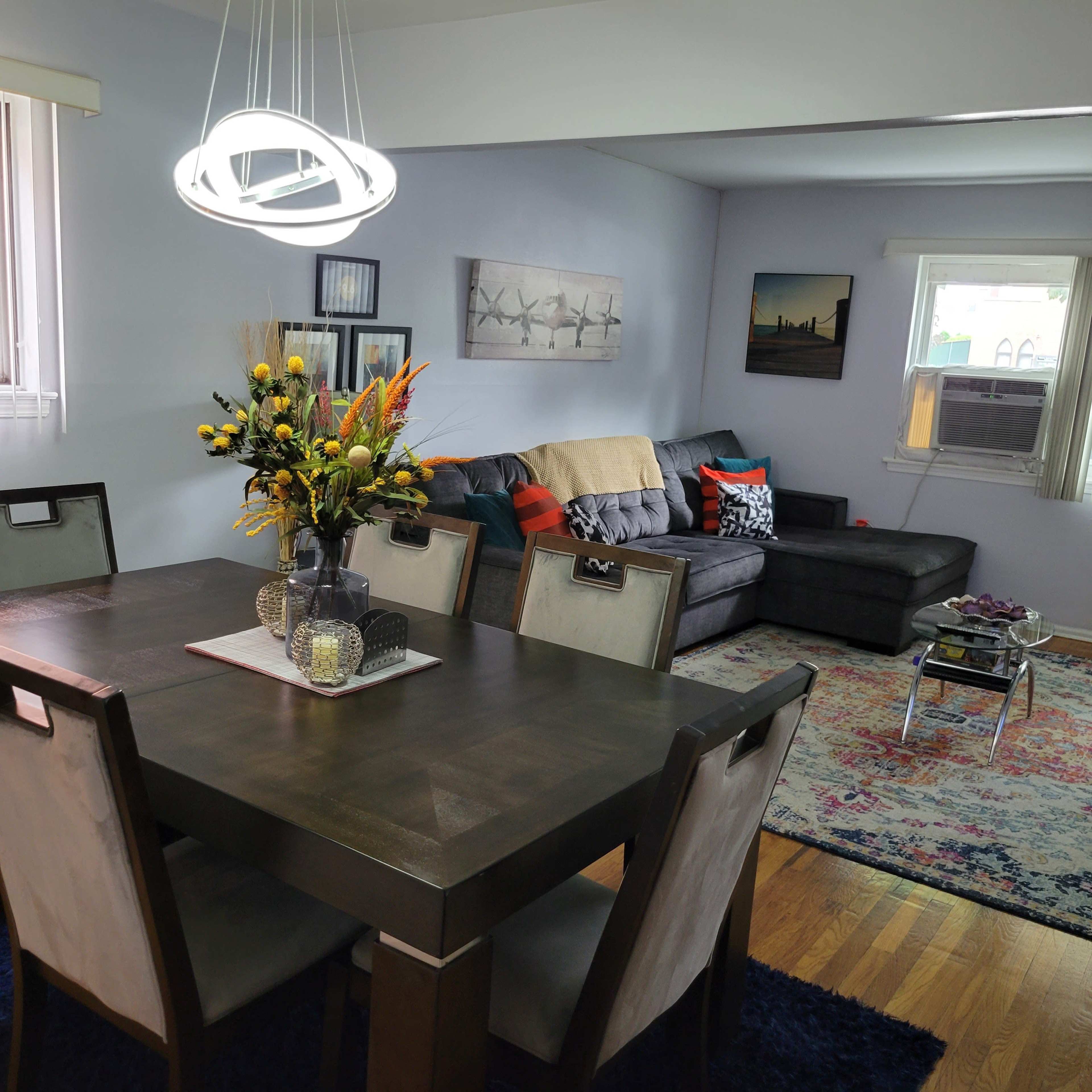 A dining area features a wooden table with chairs, a floral centerpiece, and a modern light fixture, while a living room with a gray sectional sofa and decorative pillows is visible in the background.
