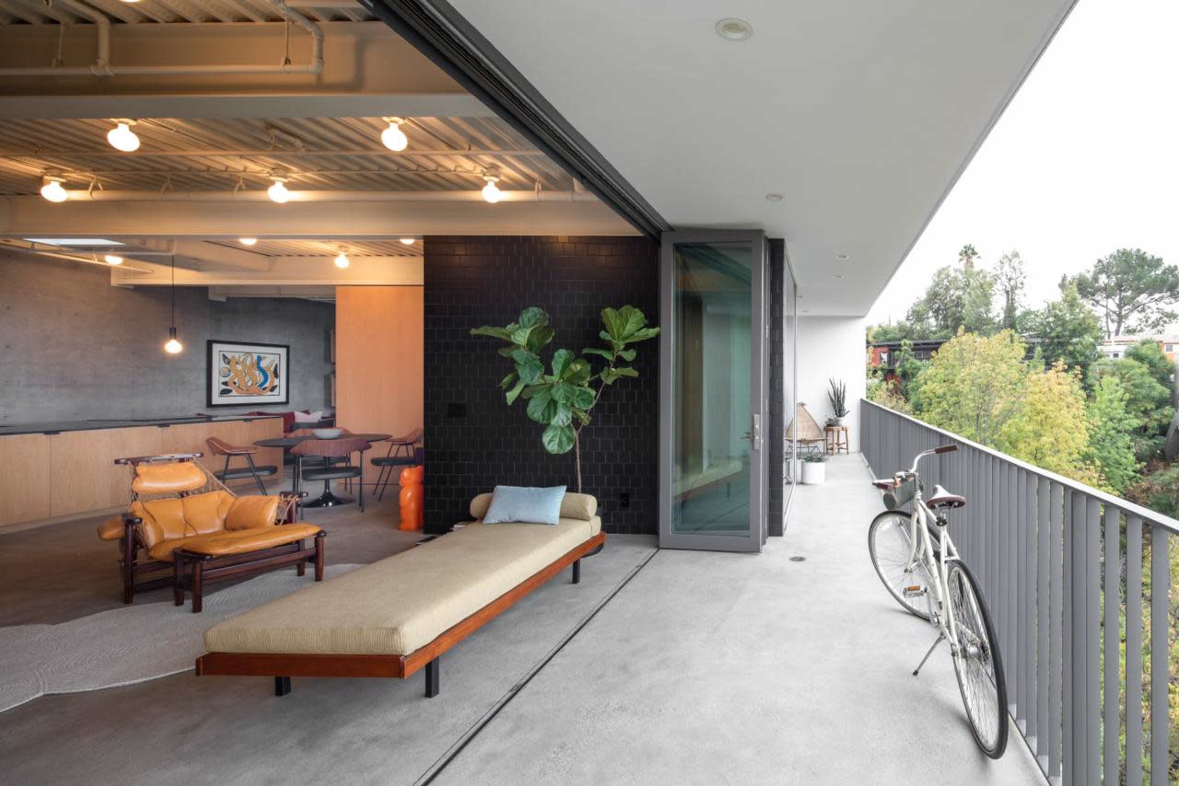 A modern balcony features a long bench, a bicycle resting against the railing, and an open doorway leading into a sleek indoor living space.
