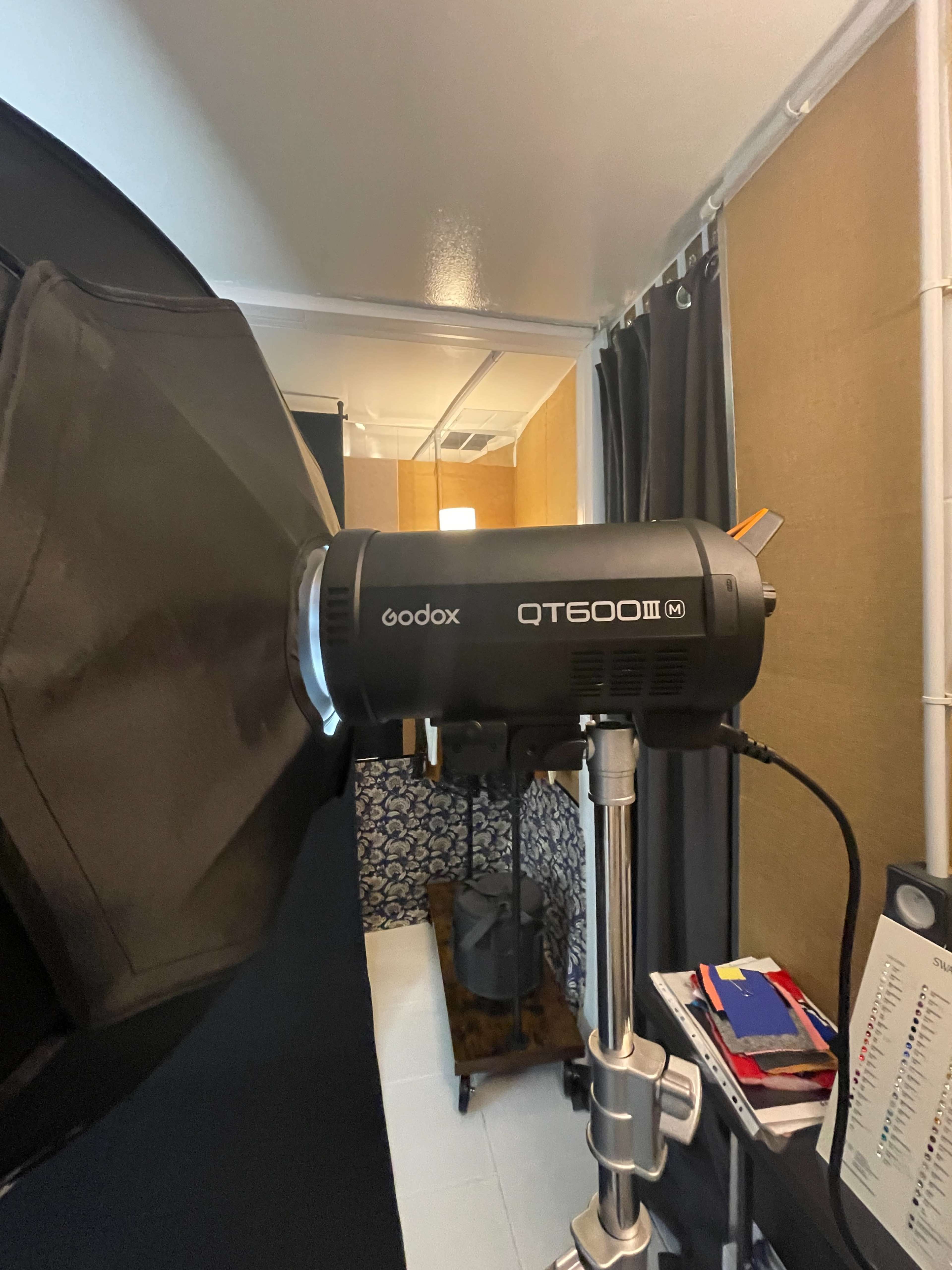 The image shows a Godox QT600 III studio flash mounted on a stand, with a background featuring a textured wall and fabric curtains.
