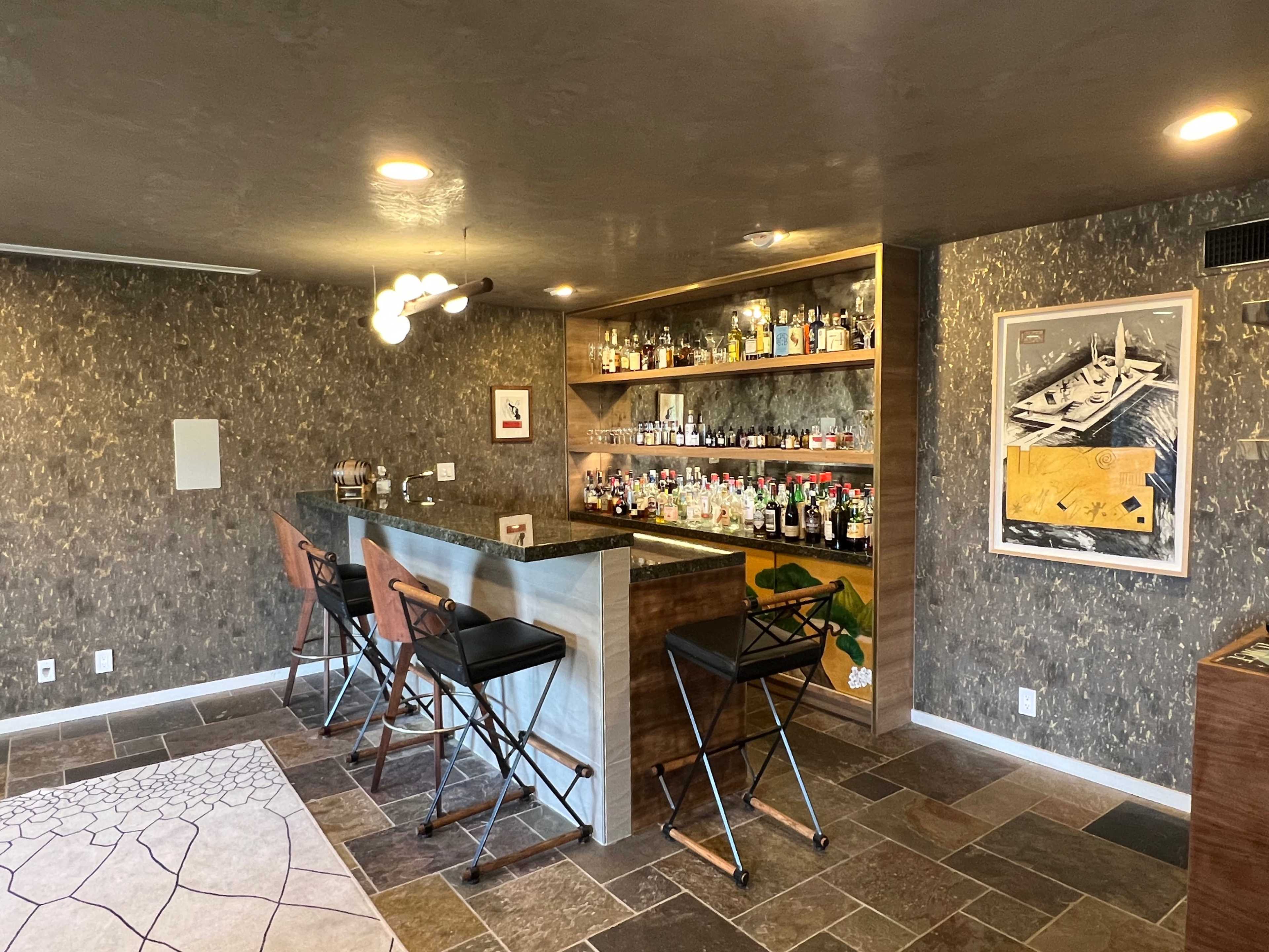 The image shows a well-organized home bar area featuring a long countertop with high stools, a display of various bottles on shelves above, and artwork on the walls.