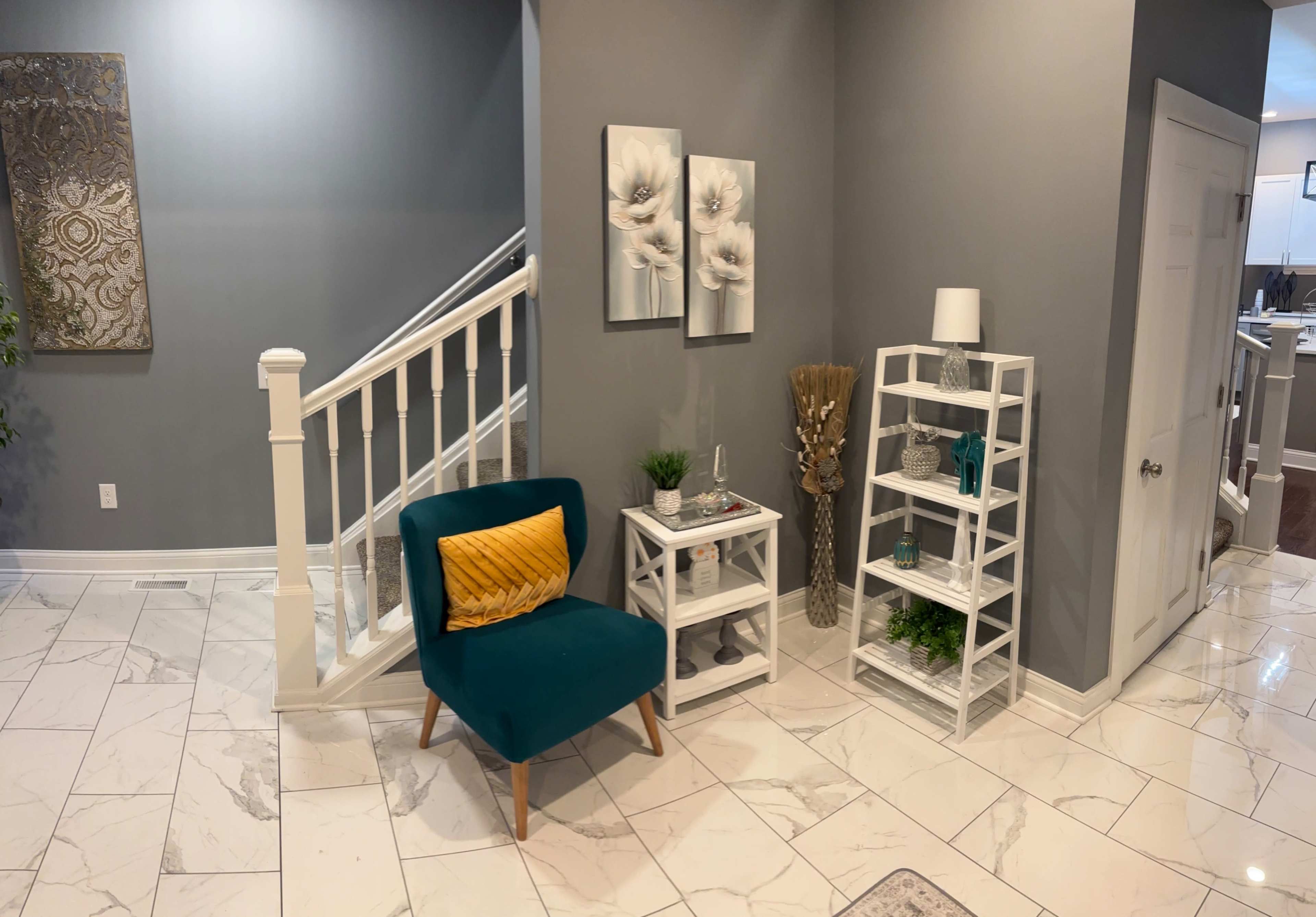 A small entryway features a teal chair with a yellow cushion, white shelving units with decorative items, and a staircase against a gray wall.