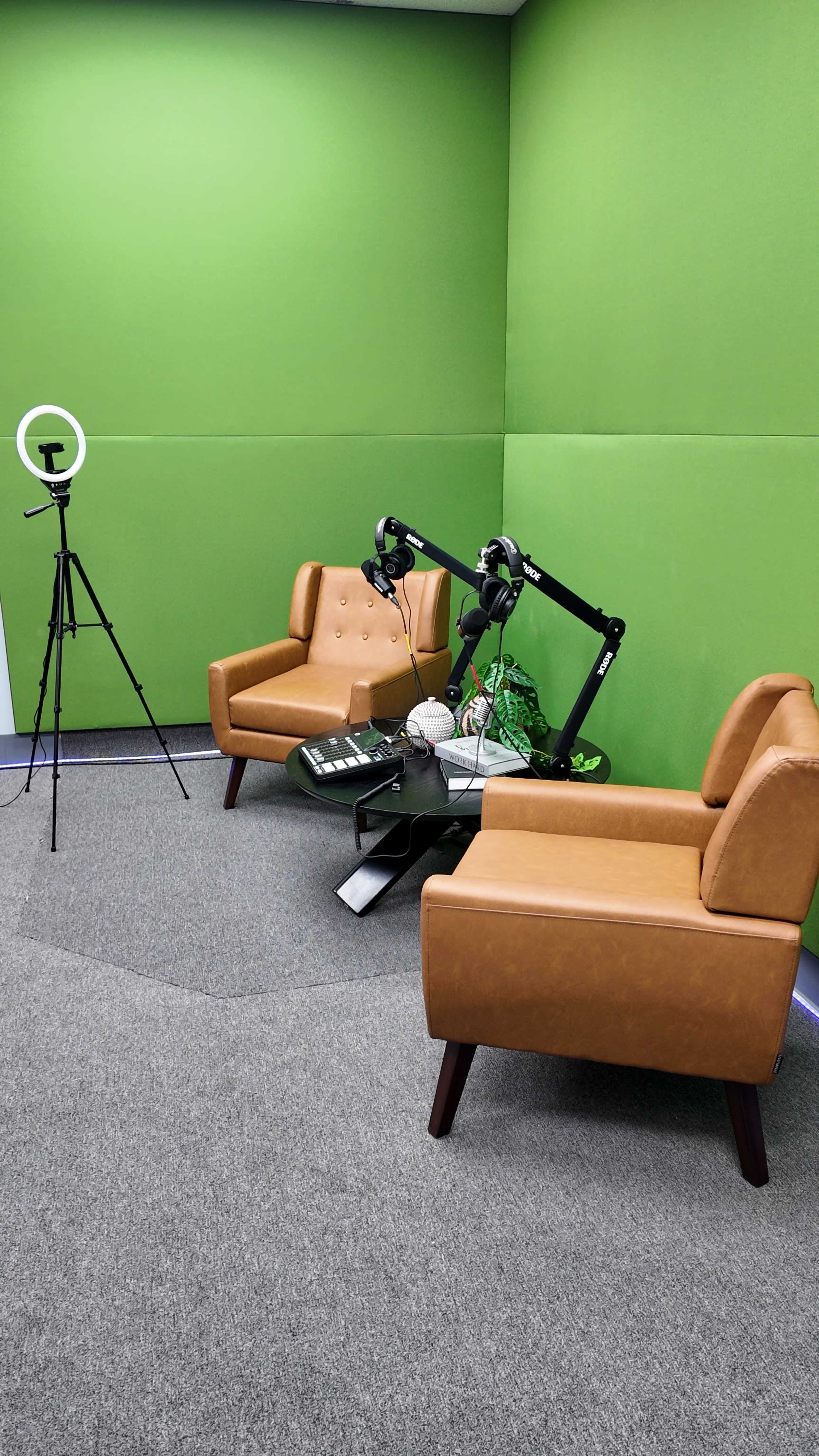 The image shows a small studio space with two tan chairs, a glass coffee table, a ring light on a tripod, and microphone stands, all set against green walls.