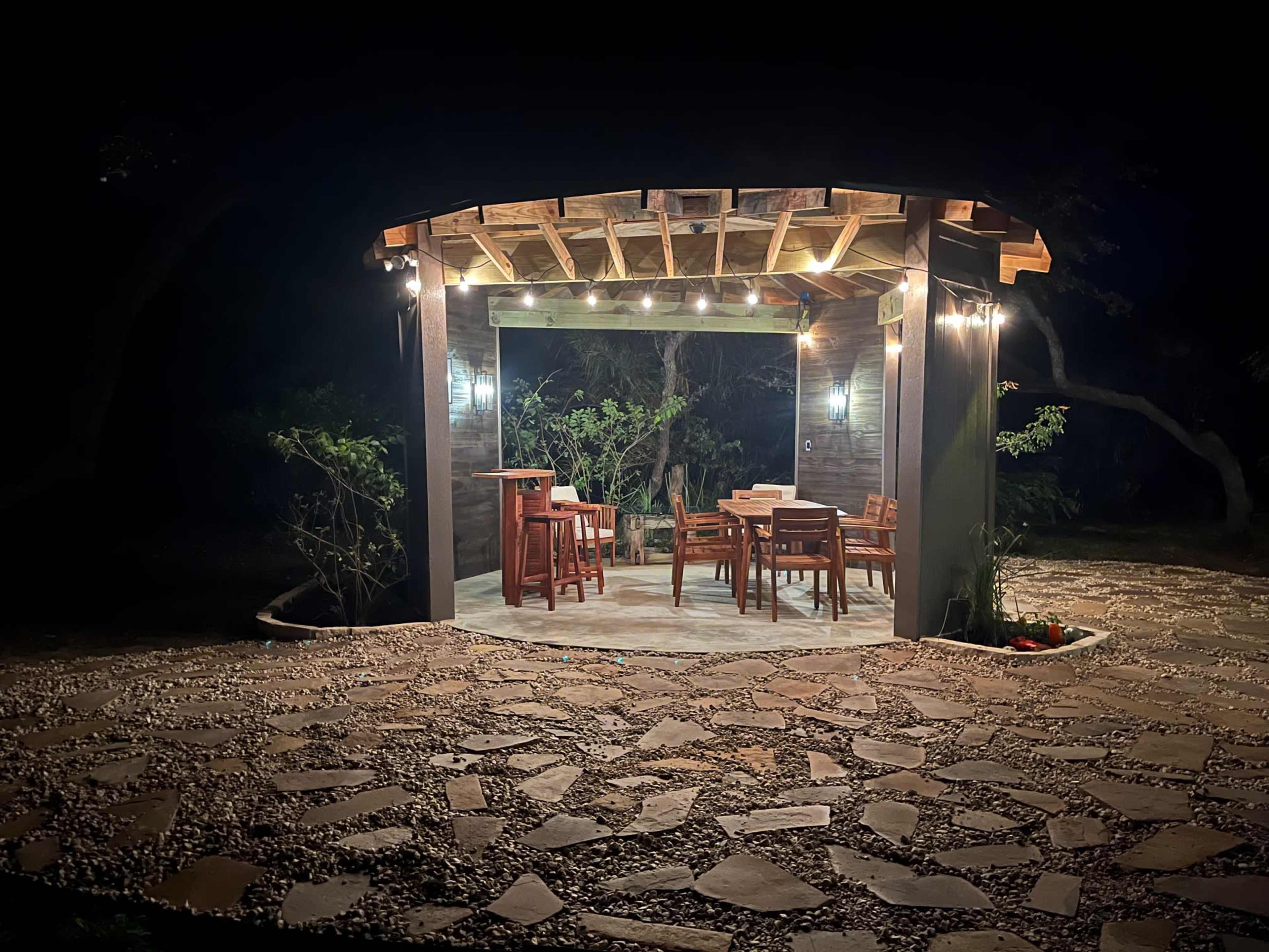A wooden gazebo with tables and chairs is illuminated by string lights, surrounded by a stone path in a dark outdoor setting.