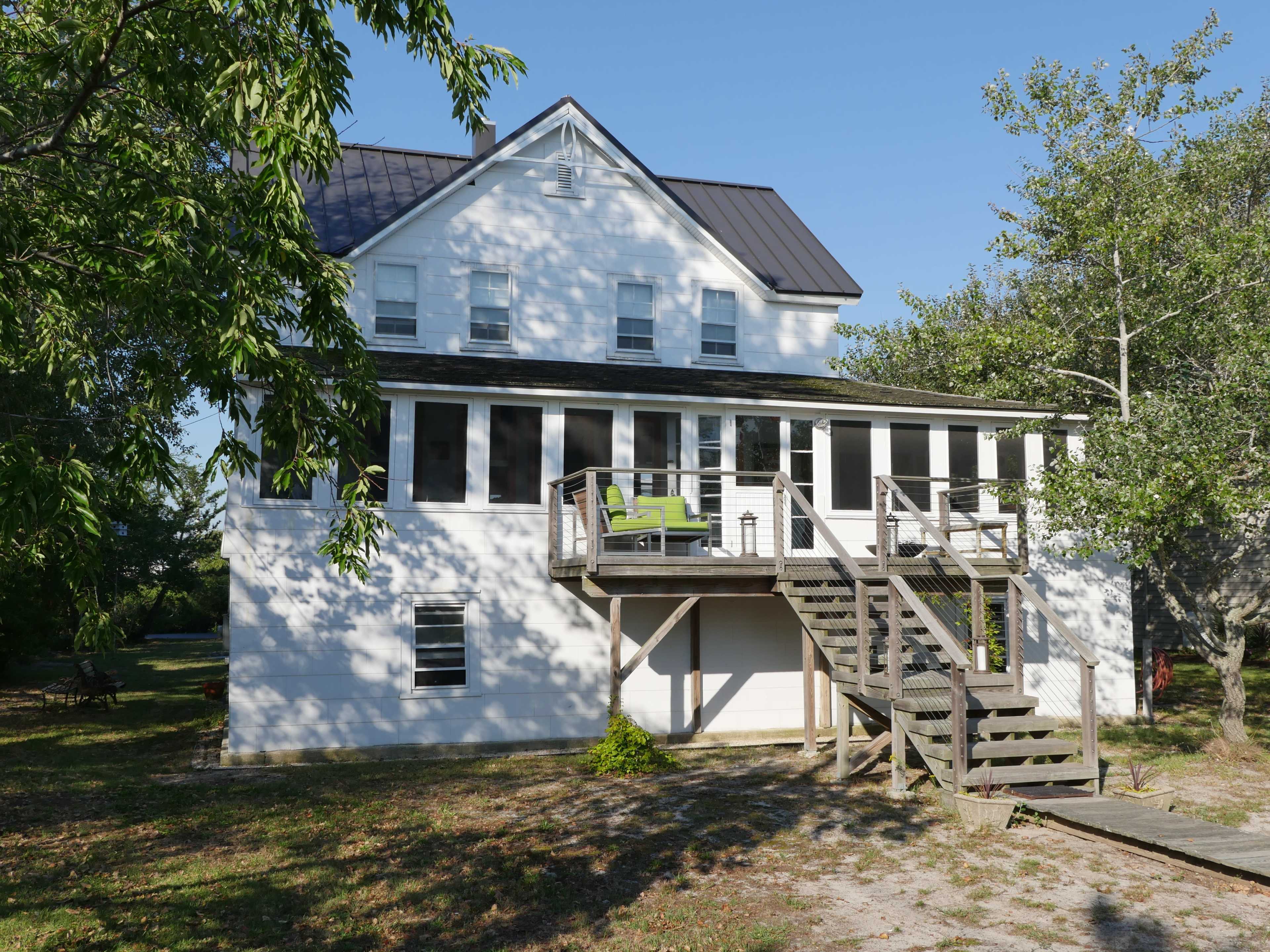 A two-story white house with a dark metal roof features a front porch and wooden stairs leading to a deck.