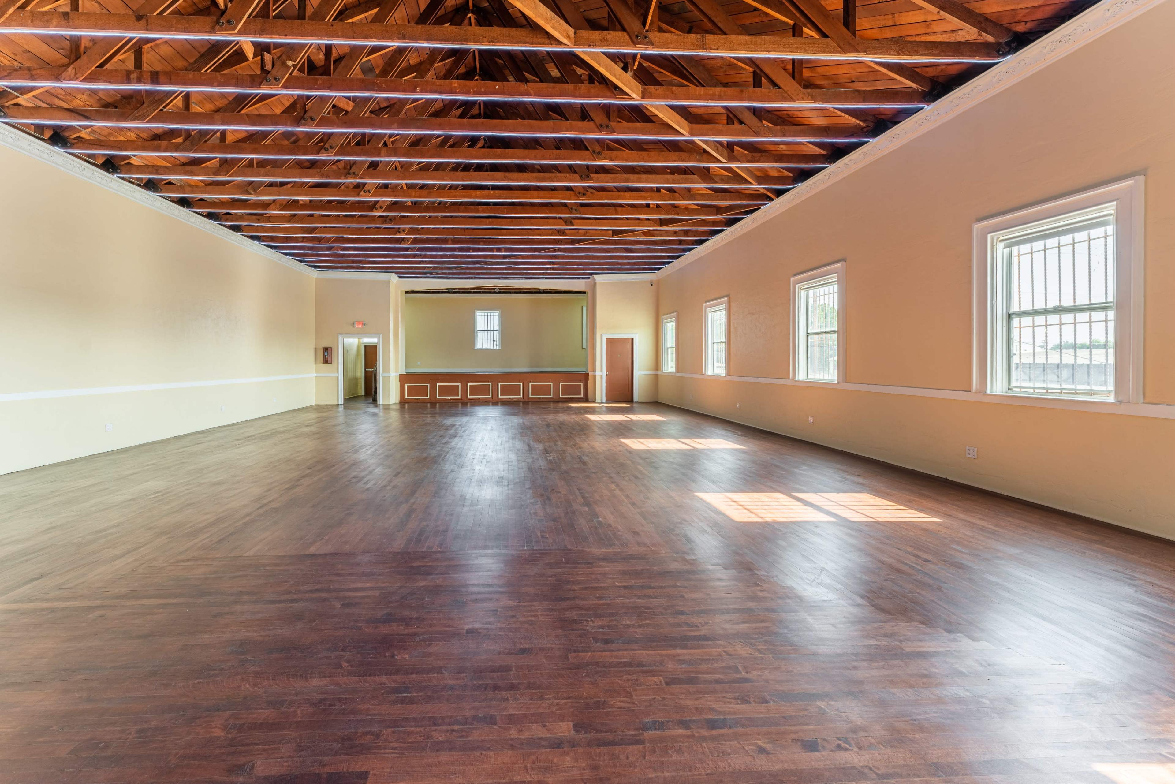 A spacious room with wooden flooring, high ceilings supported by wooden beams, and large windows allowing natural light to enter.