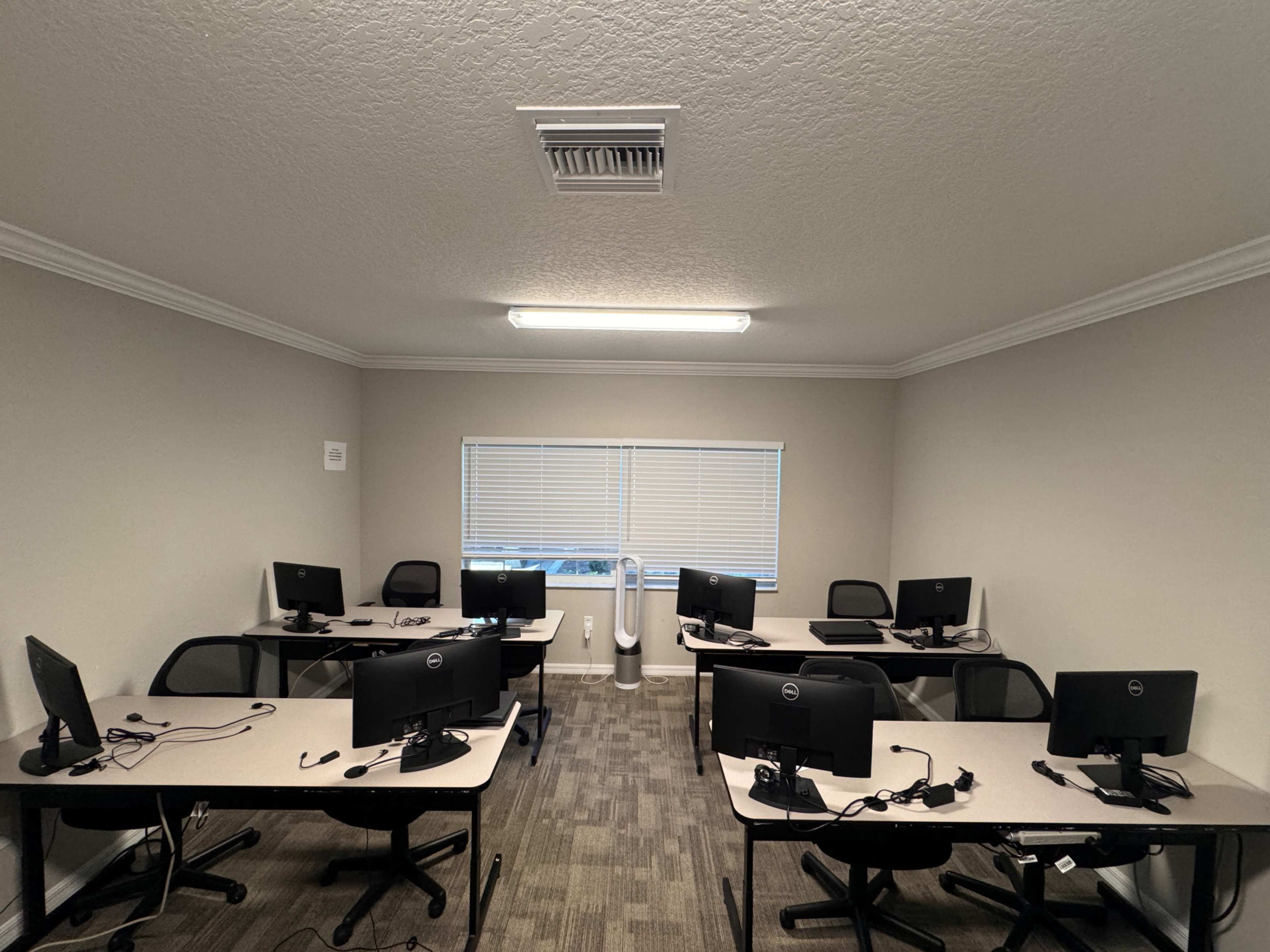 The image shows a classroom or office space with several desks, each equipped with a computer and chair, arranged neatly in a well-lit room.