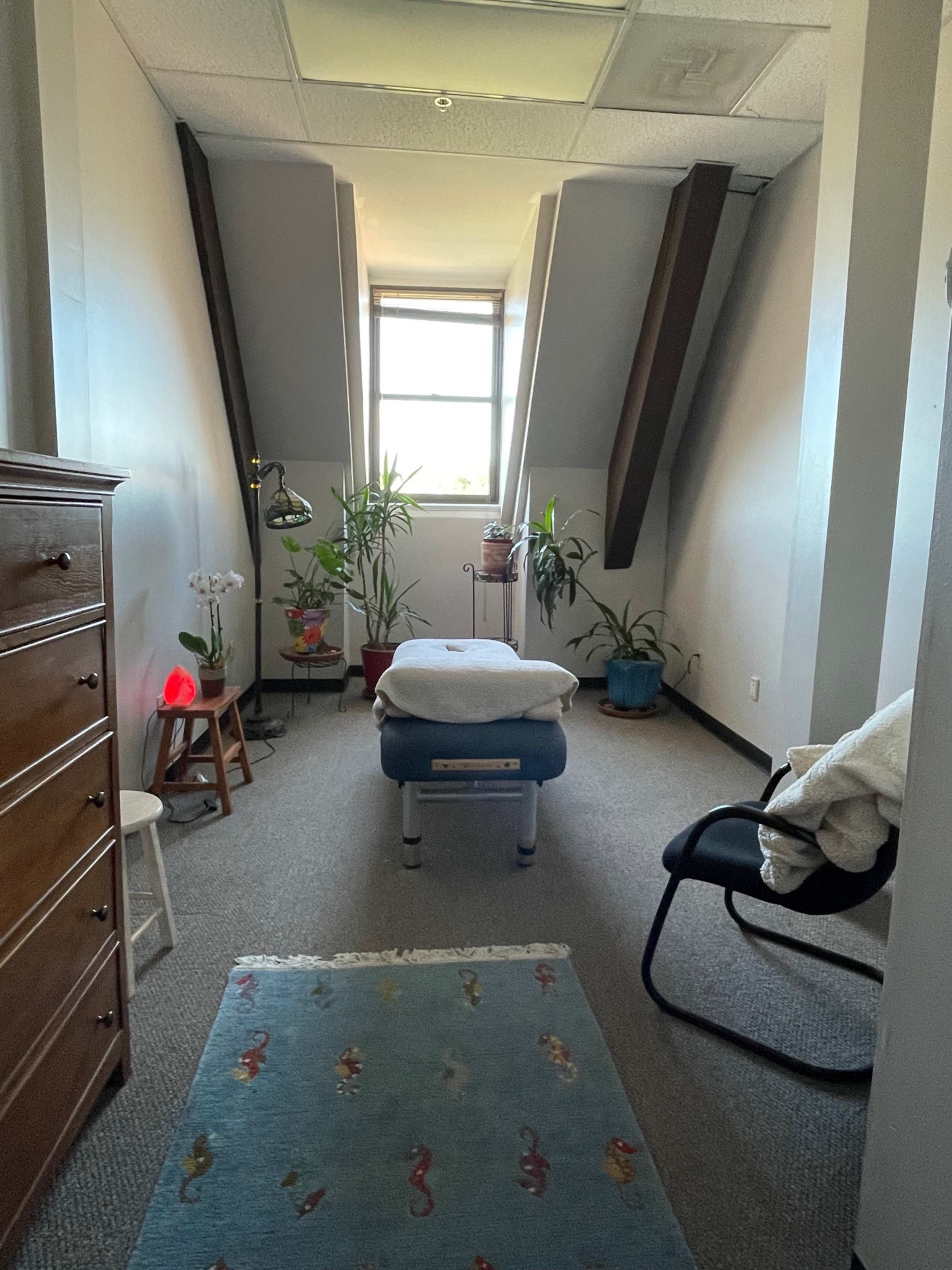 A therapy room with a massage table in the center, flanked by potted plants, a chair, a dresser, and a window providing natural light.
