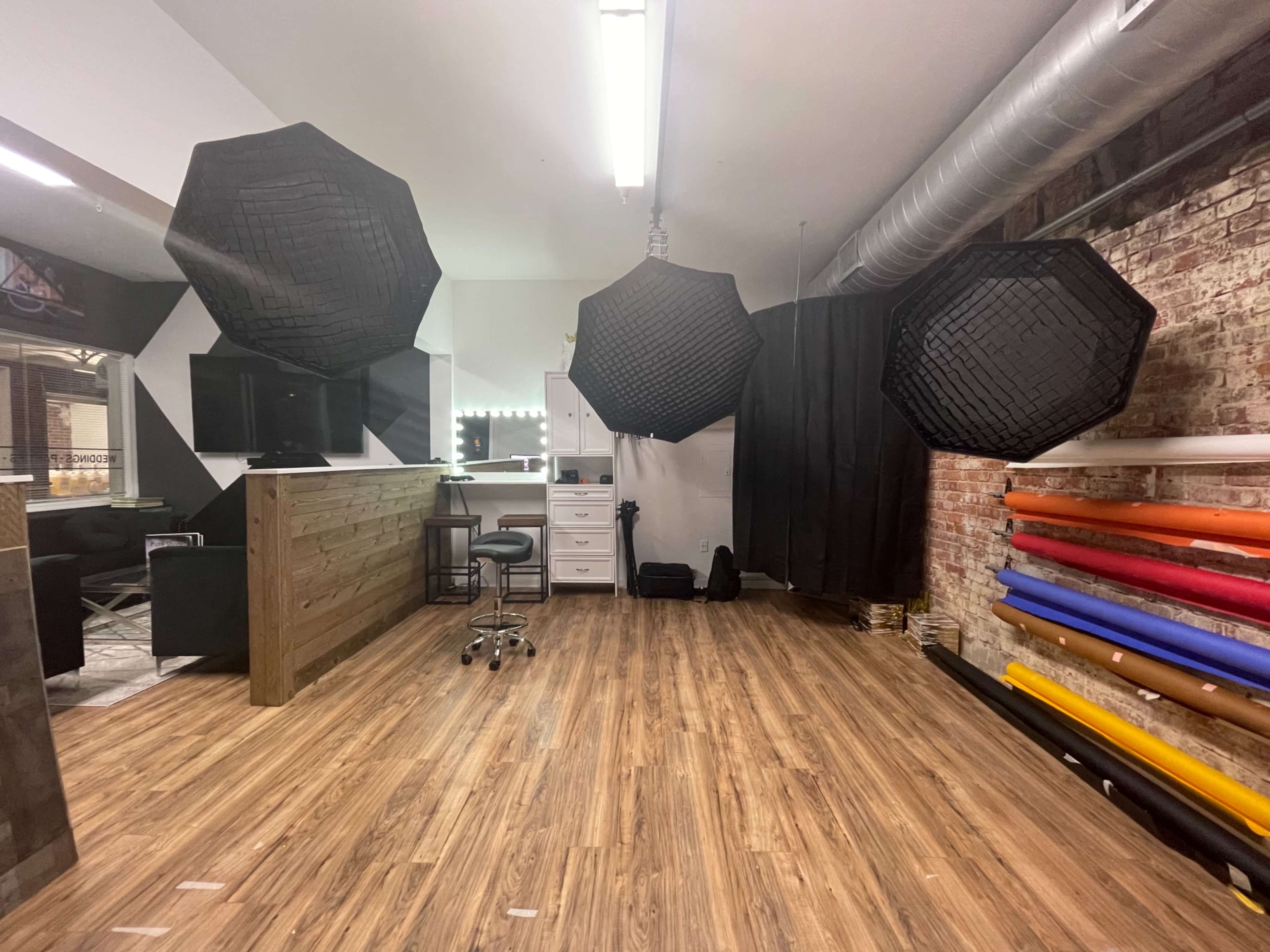 A photography studio with three large softboxes hanging overhead, a wooden reception desk, a chair, and rolls of colored backdrops along the wall.