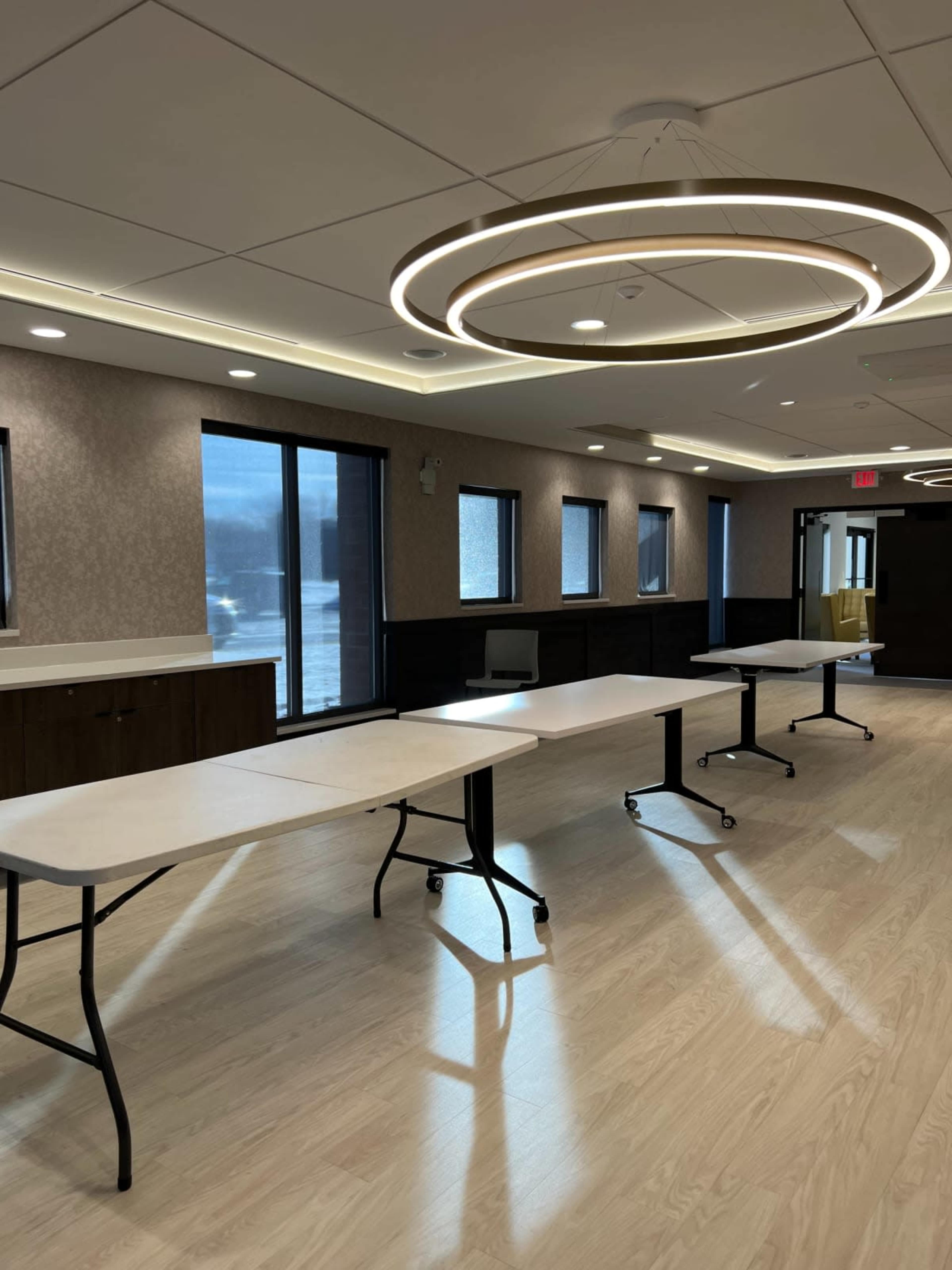 A brightly lit room with several long folding tables arranged in a row and large windows along one wall.