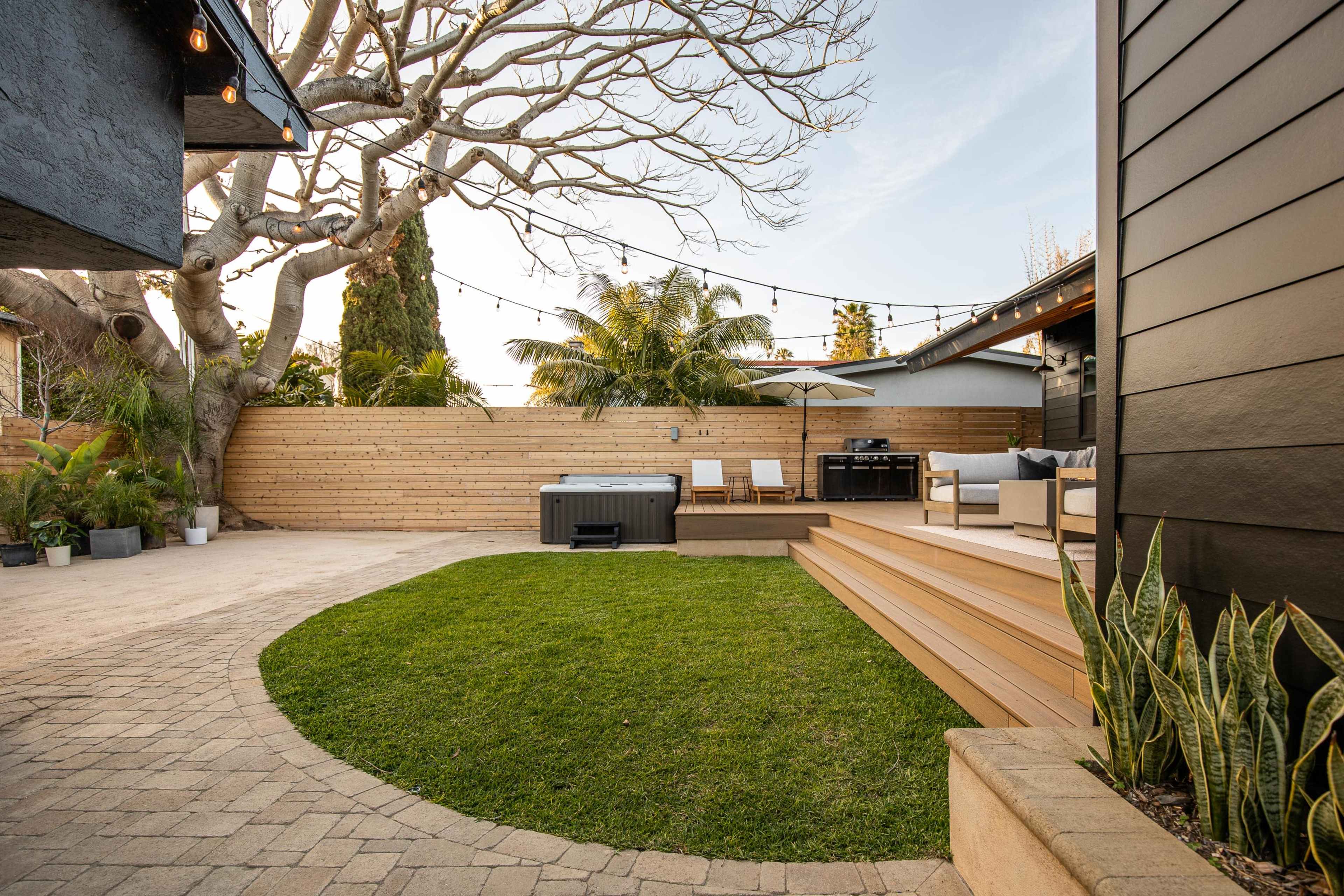 A backyard with a hot tub, seating area, and landscaped lawn surrounded by wooden fencing and a large tree.