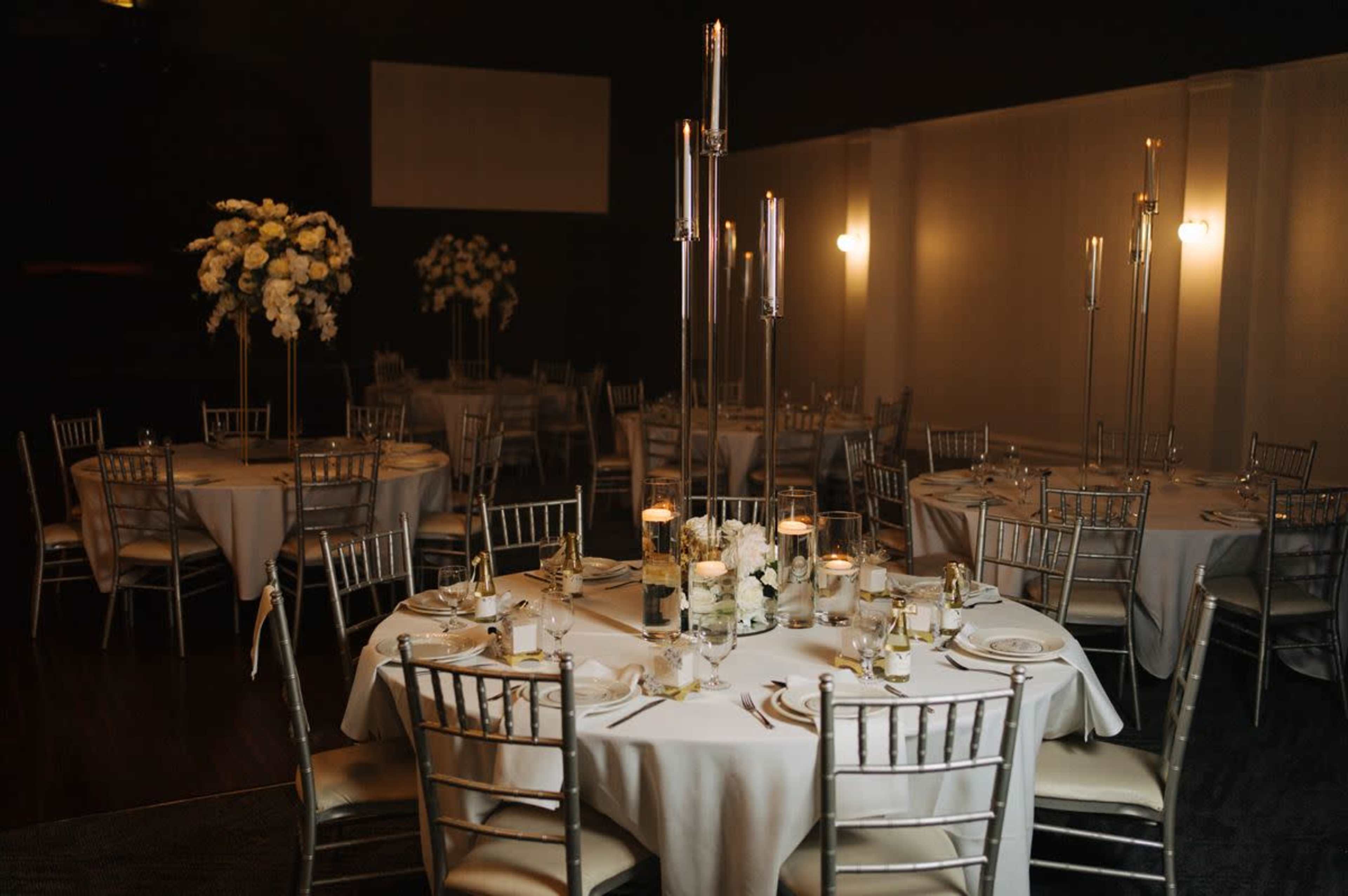 A banquet hall with several round tables set for dining, featuring silver chairs, floral centerpieces, and tall candle stands.