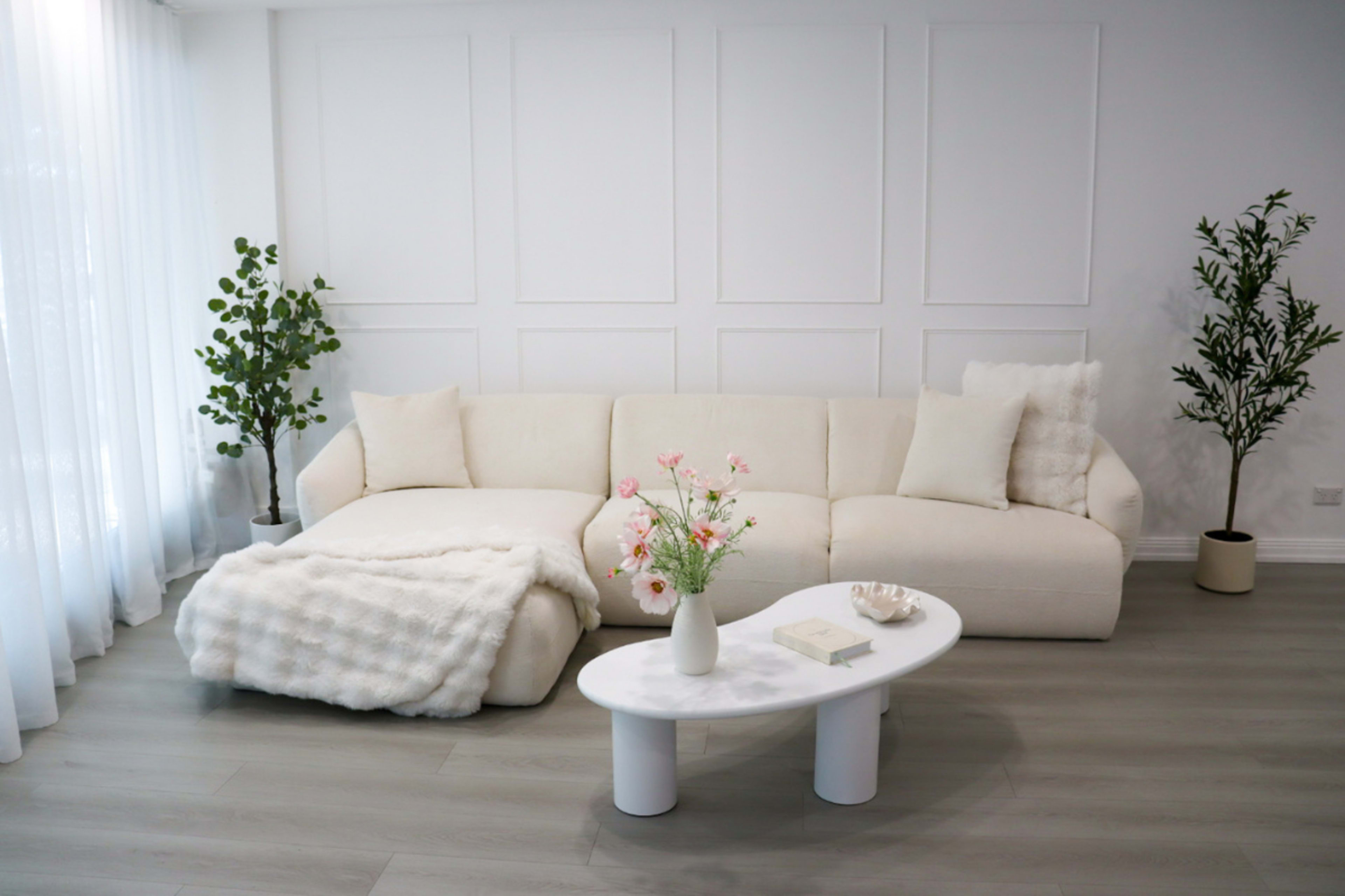 The image shows a modern living room featuring a white sectional sofa with decorative pillows, a small white coffee table, and two indoor plants positioned on either side of the sofa.
