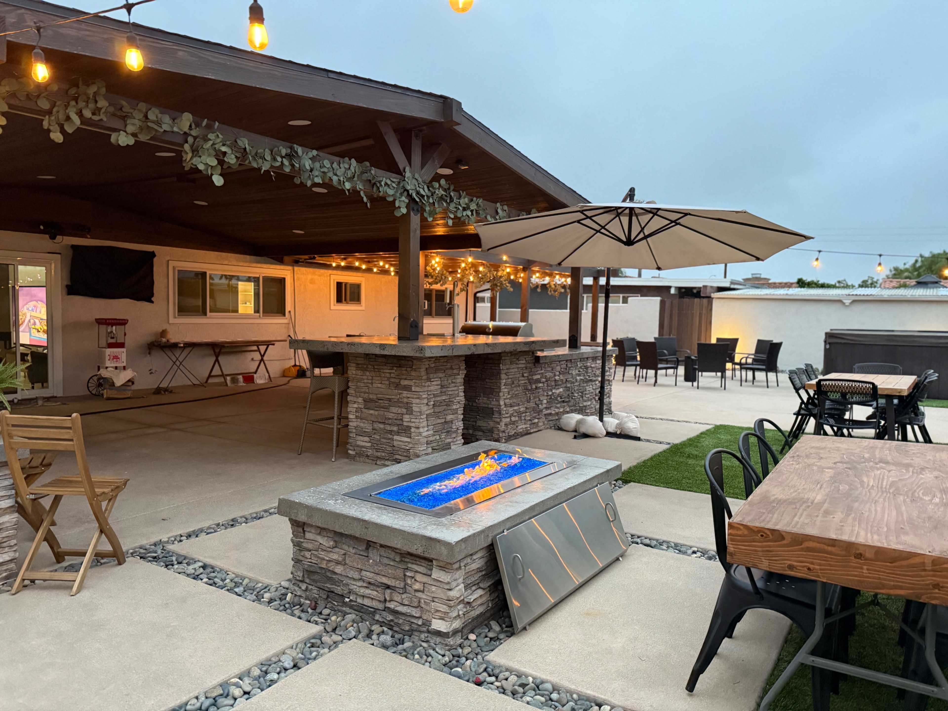 The image shows a landscaped outdoor patio area featuring a stone fire pit, dining tables, an umbrella, and string lights overhead.