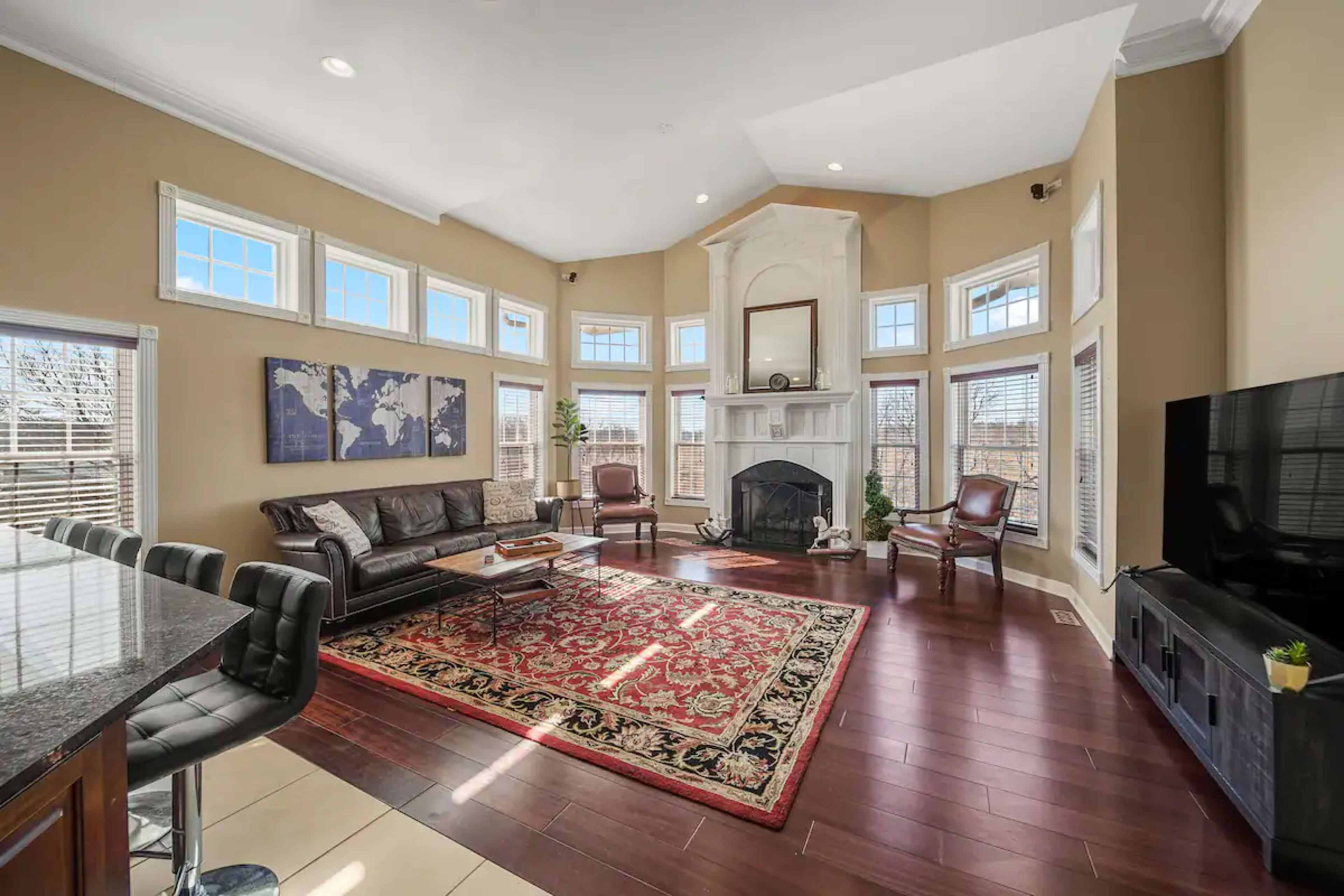 A spacious living room features a large bay window, a fireplace, and a patterned area rug, with seating arranged around a coffee table.