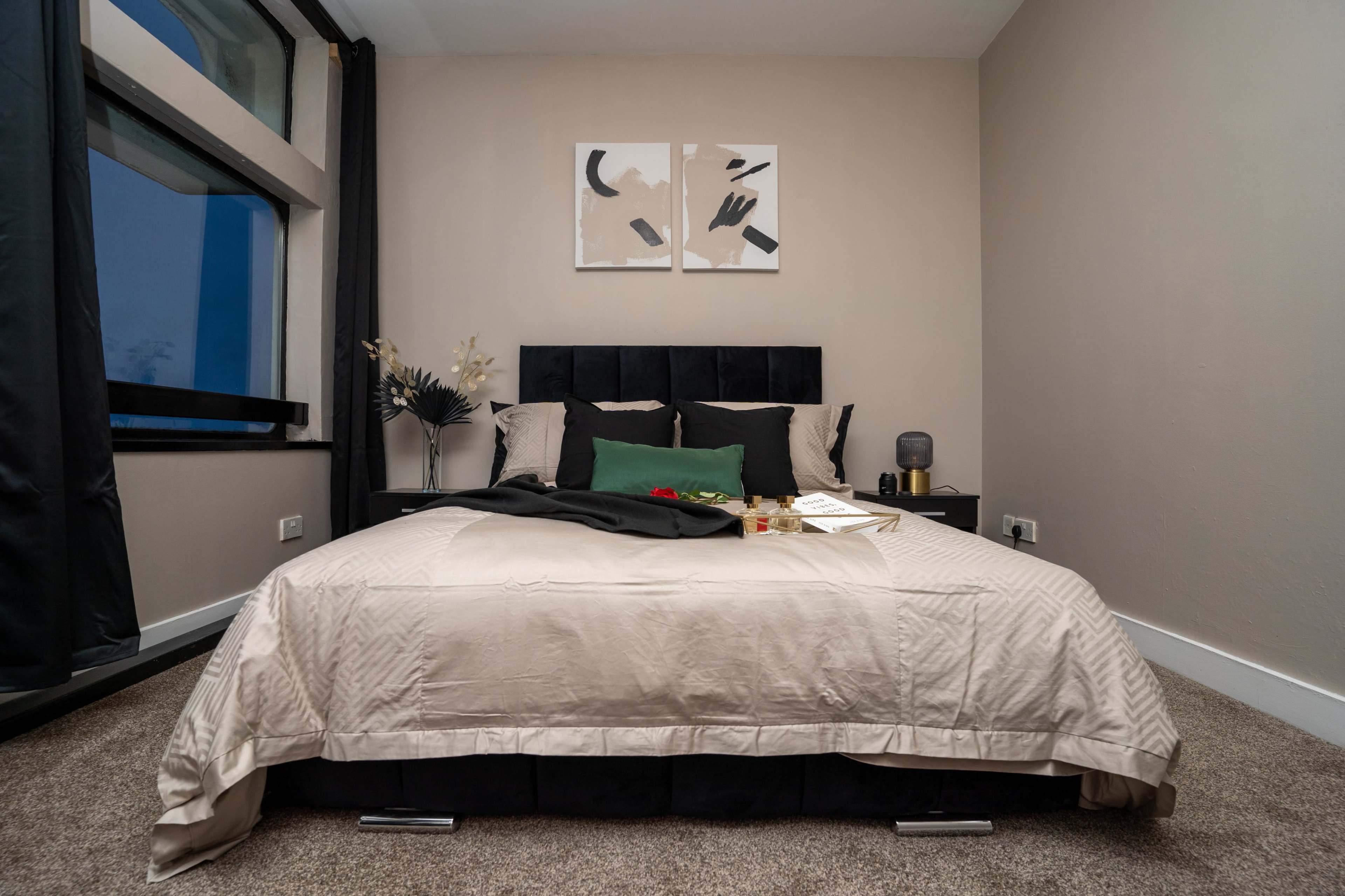 A neatly made bed with a light-colored comforter and decorative pillows in a modern bedroom featuring large windows and minimalist wall art.