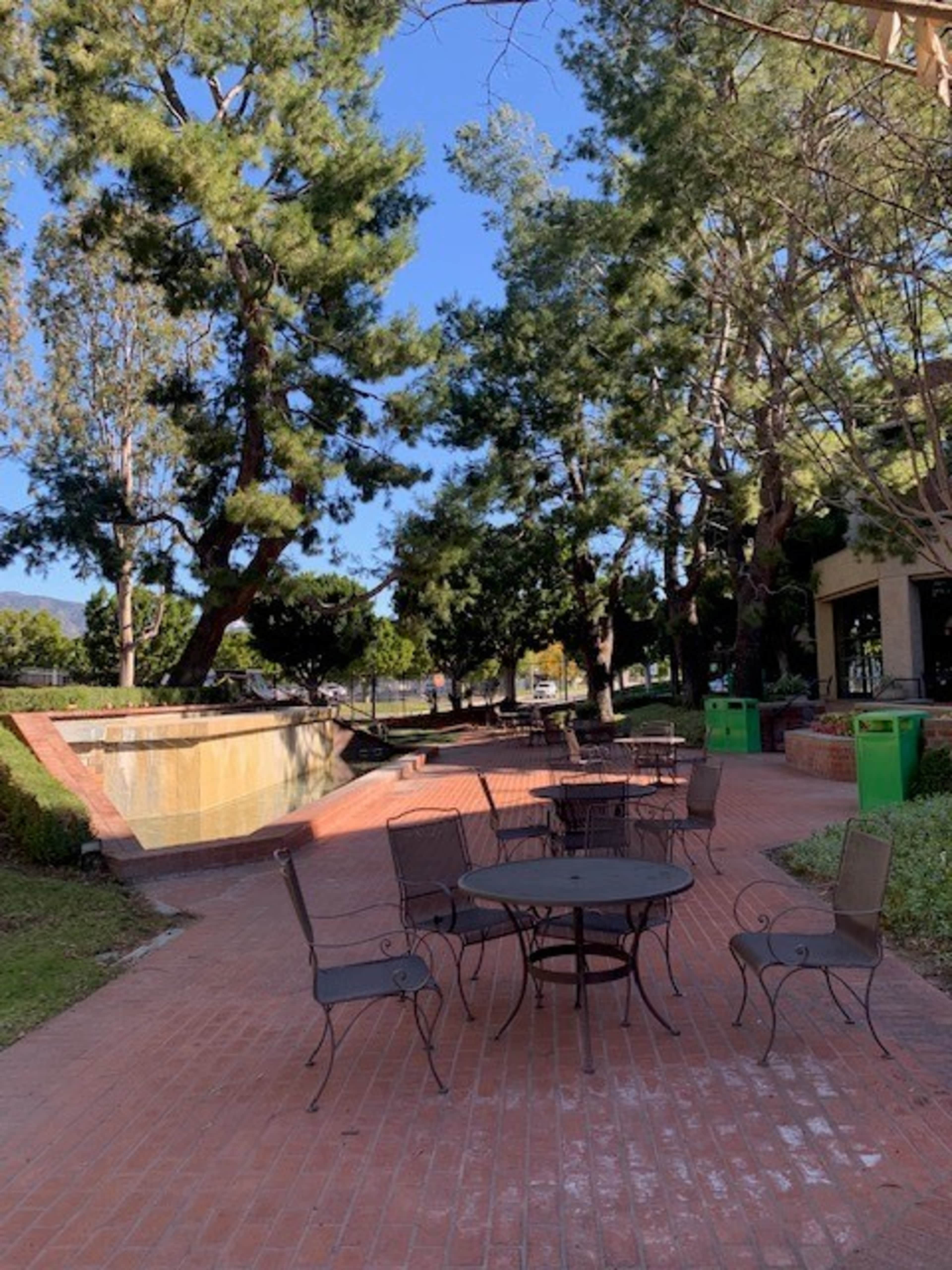 A brick walkway lined with small metal tables and chairs, surrounded by trees and leading to a water feature.