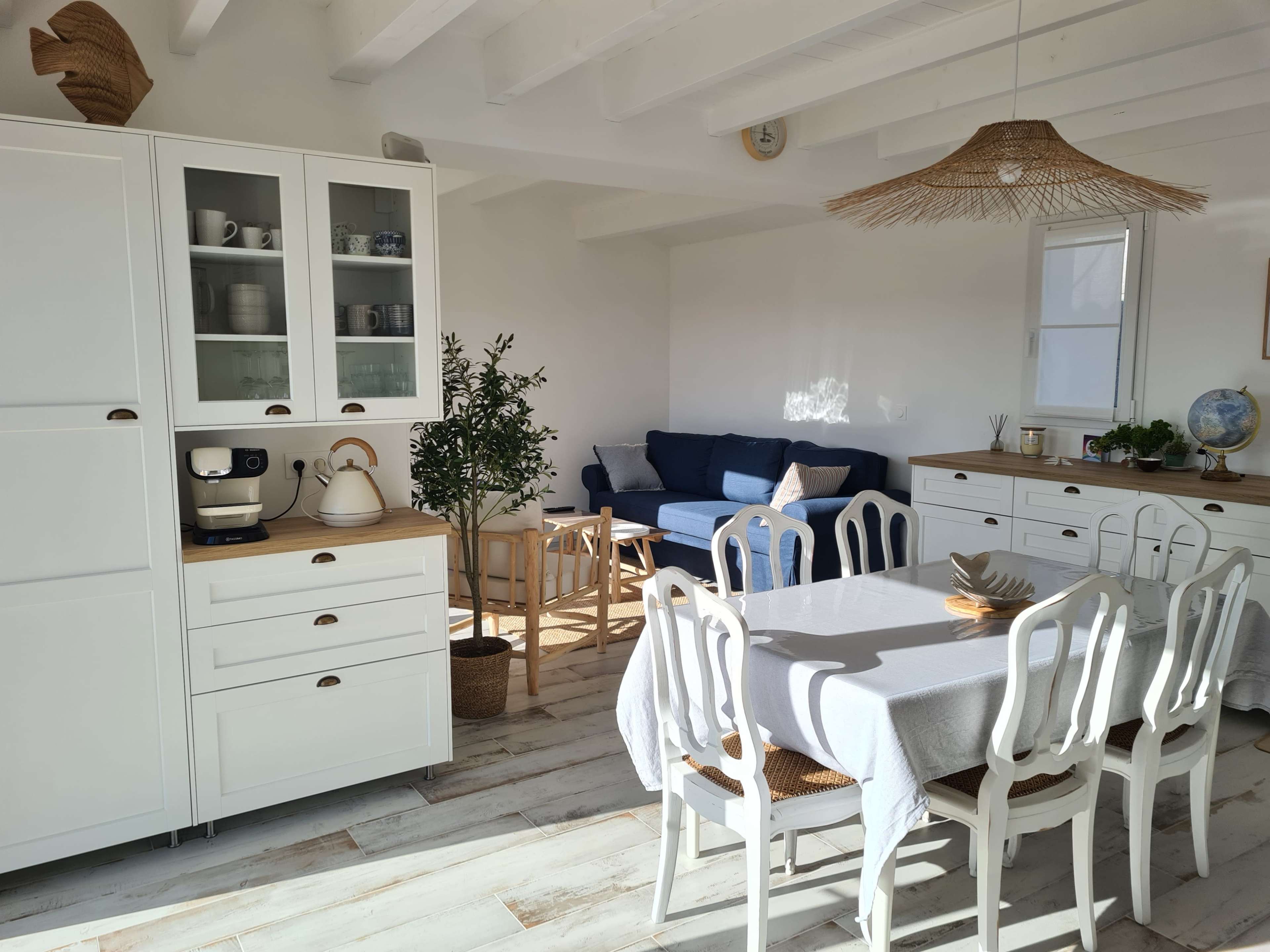 The image shows a bright, modern kitchen and living area featuring a white table with chairs, a sleek kitchen cabinet, and a cozy seating area with a blue sofa.