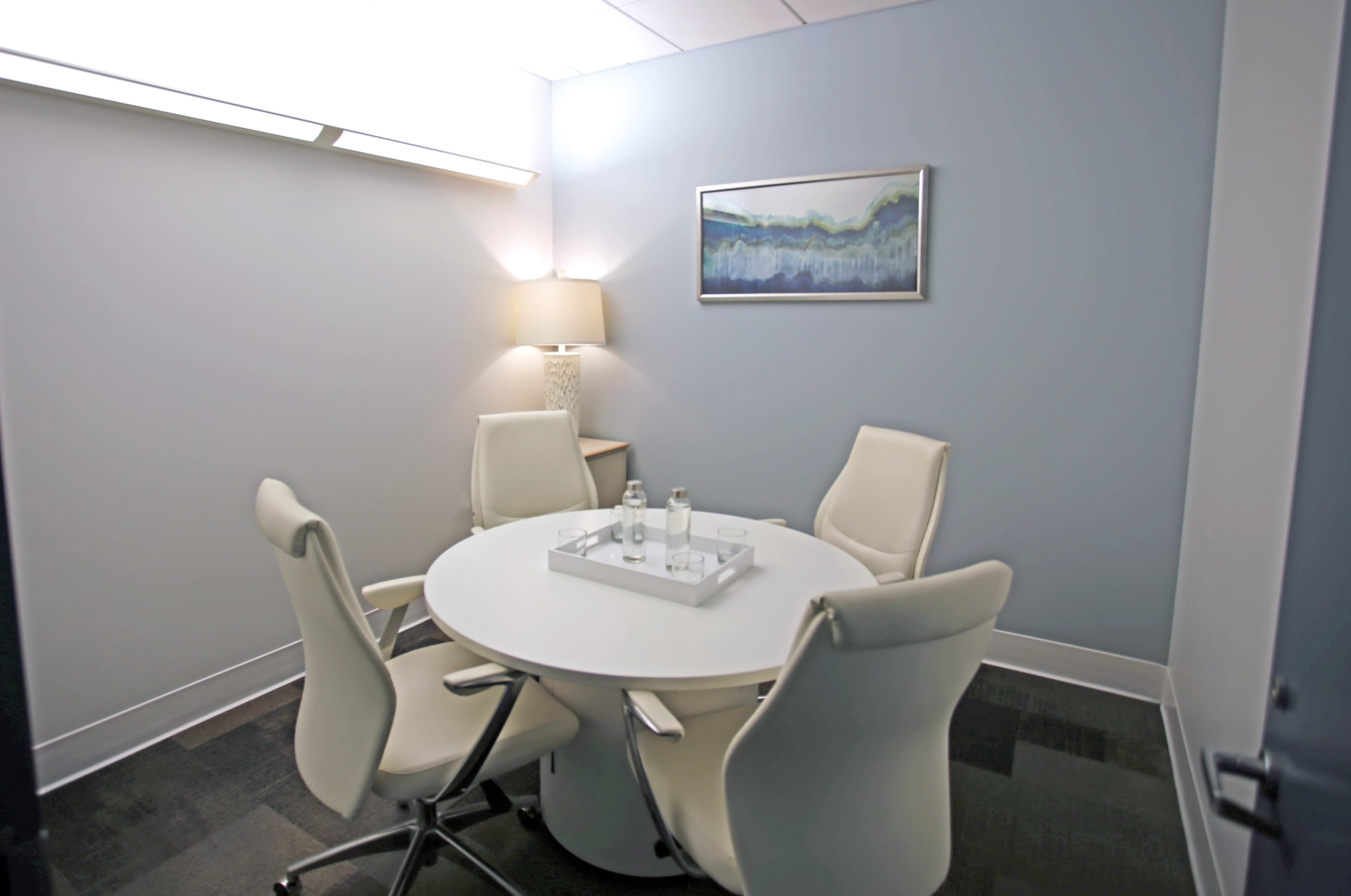 A small meeting room features a round table surrounded by four white chairs, with a lamp and a framed artwork on the walls.