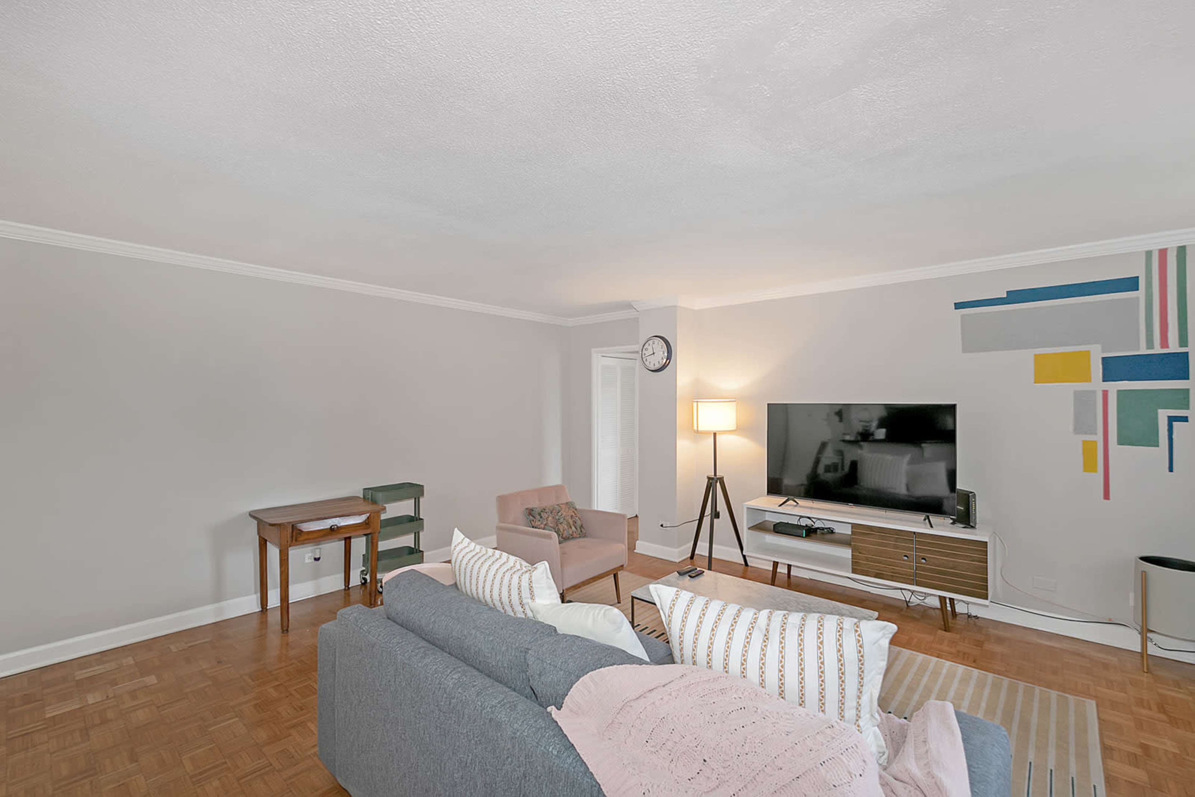 The living room features a gray sofa, a light pink chair, a TV stand with a television, and a small table against a wall with colorful geometric artwork.