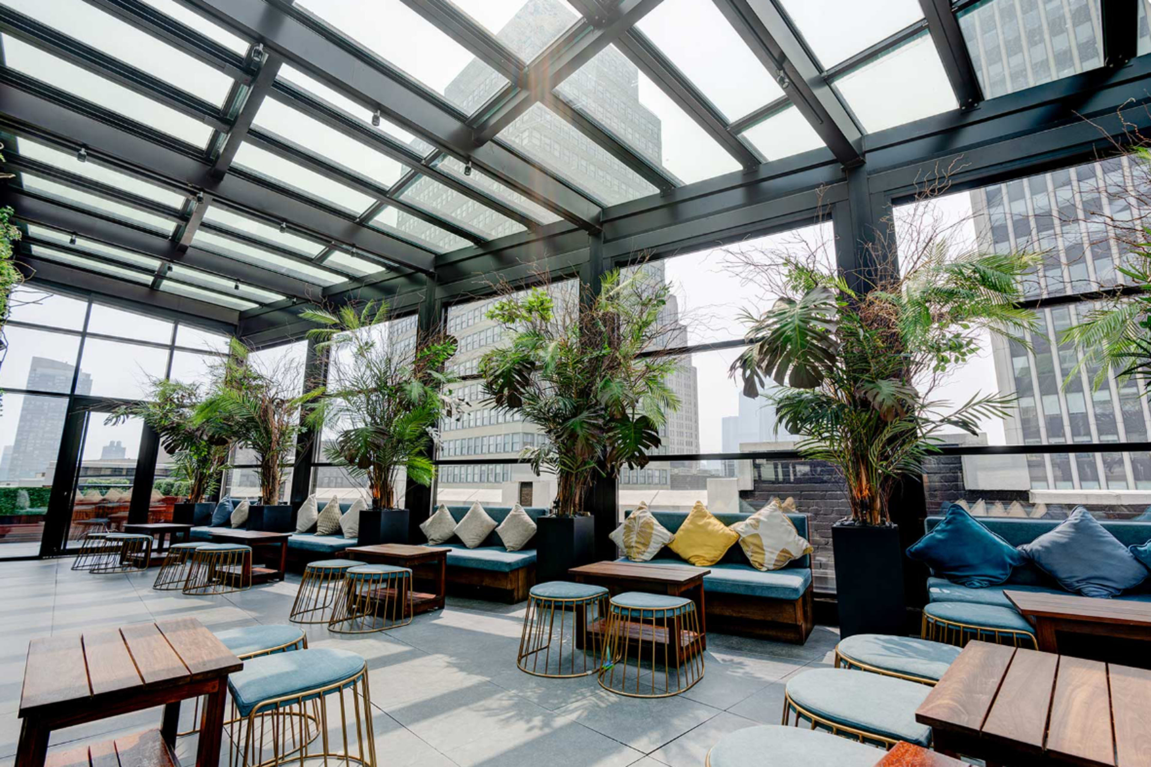The image shows a rooftop lounge featuring glass walls, potted plants, and seating areas with tables overlooking a cityscape.