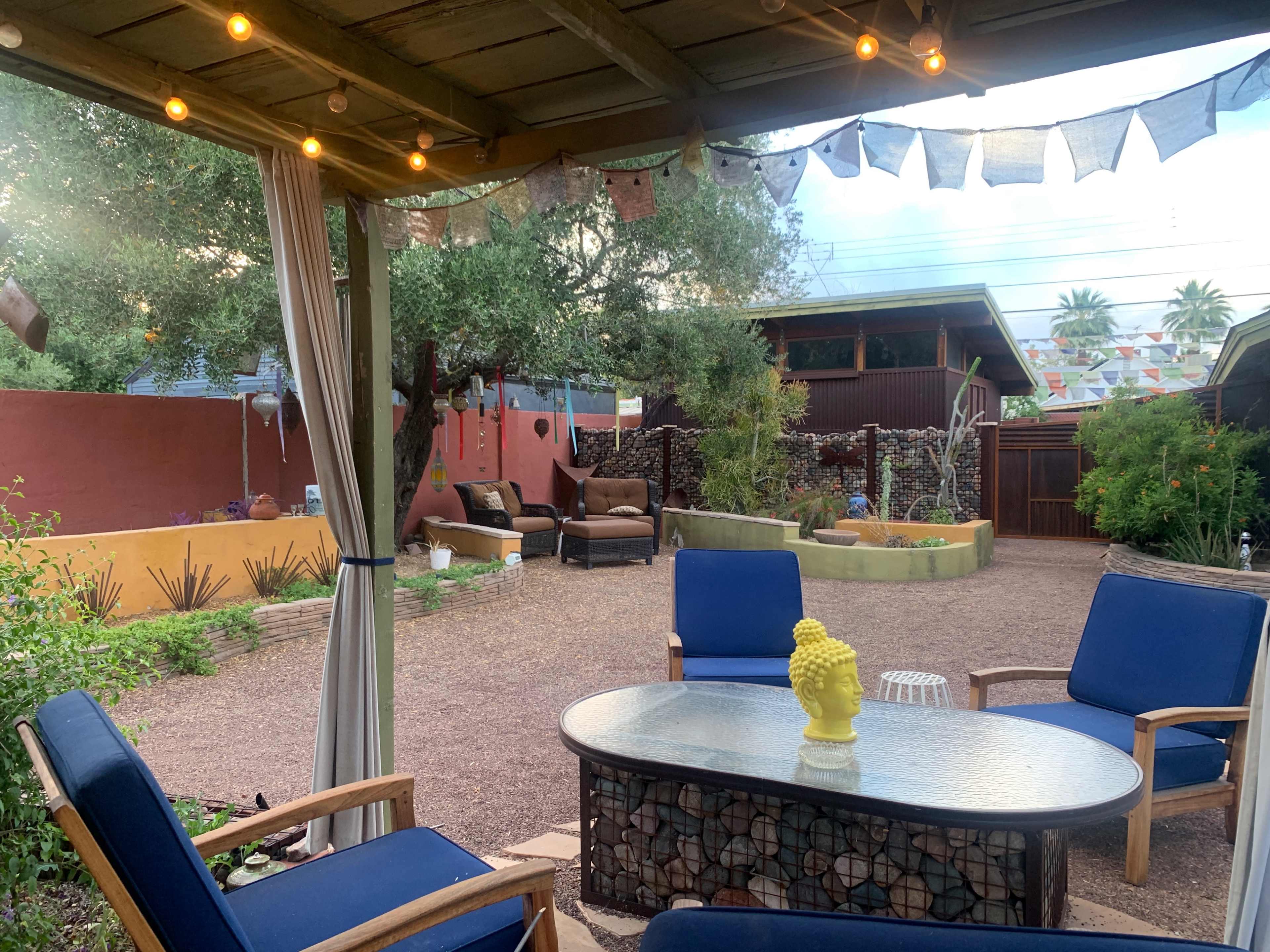 A backyard scene features a seating area with blue chairs and a glass table under a shaded pergola, surrounded by greenery and colorful decorations.