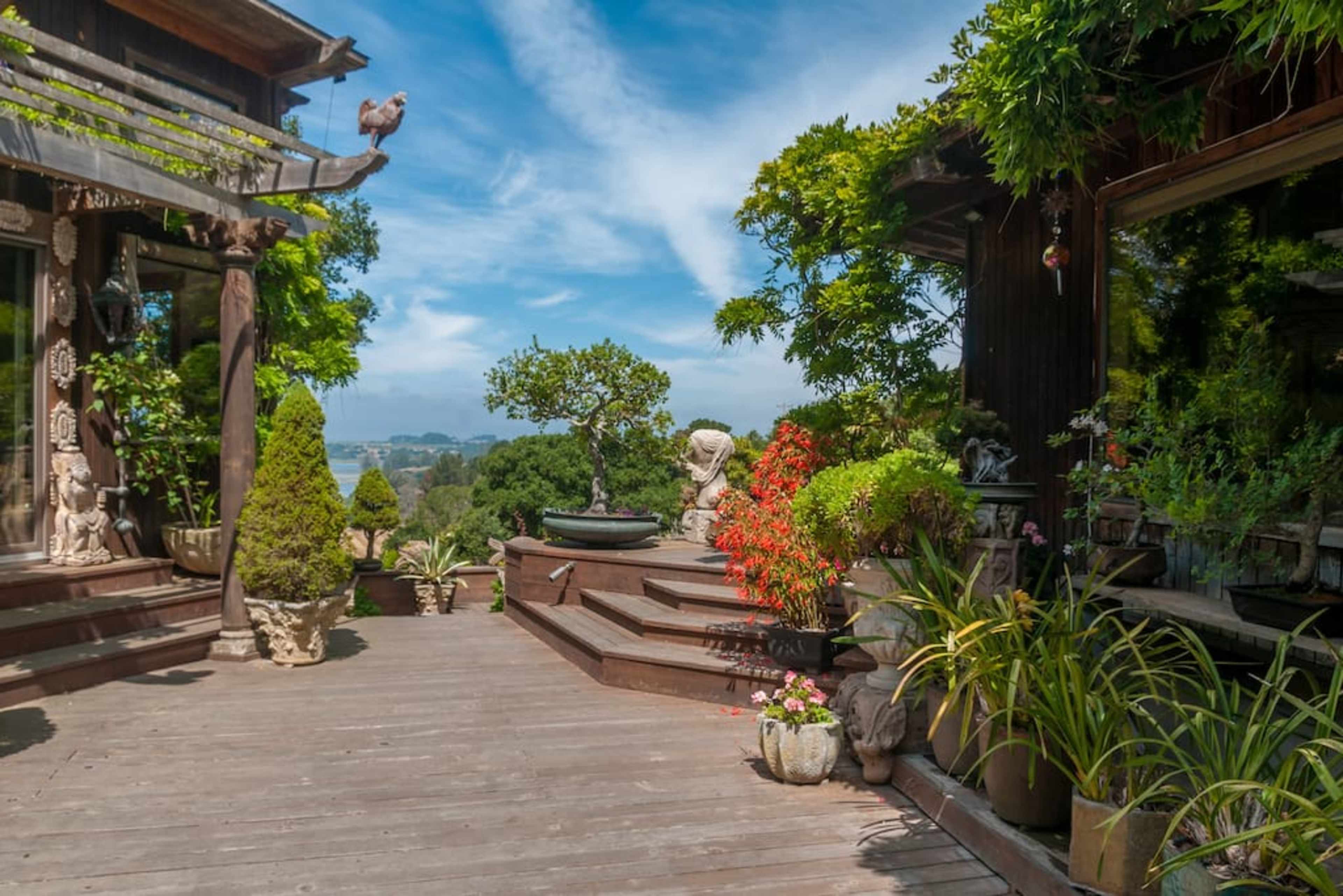 A wooden deck surrounded by lush greenery, decorative plants, and a stone statue, with a view of distant hills under a blue sky.