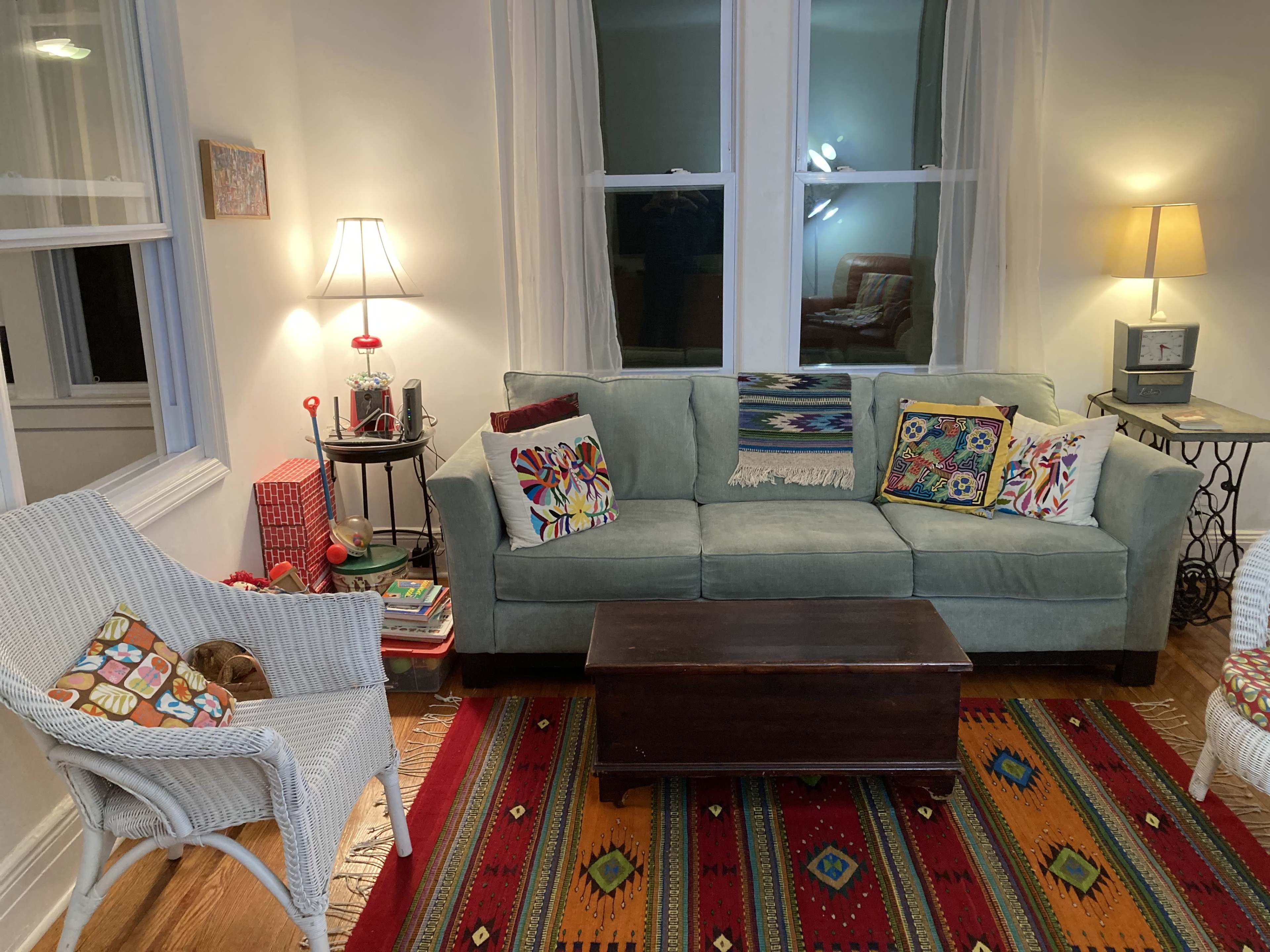 The living room features a green couch with decorative pillows, a wooden coffee table, and a white wicker chair on a patterned rug, illuminated by two lamps.