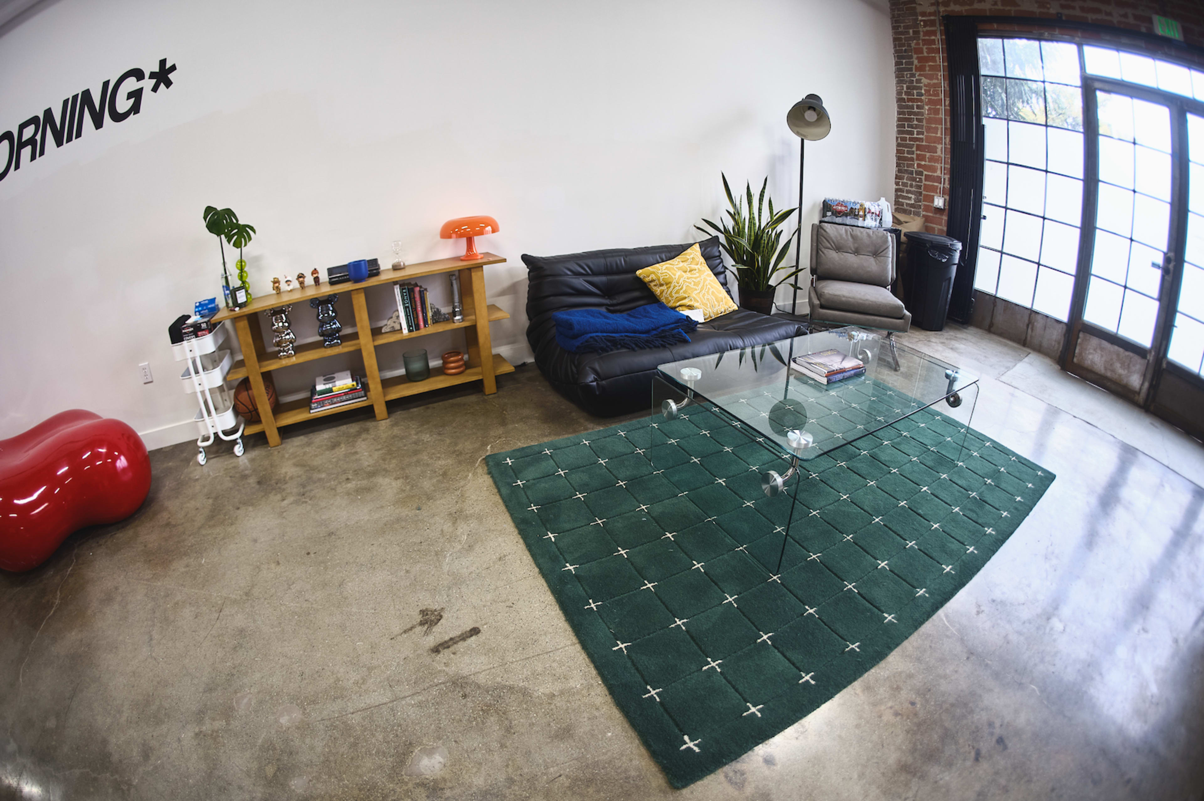 The scene depicts a modern living room featuring a black leather sofa, a glass coffee table, a green area rug with a geometric pattern, and a wooden shelf filled with books and decorative items.