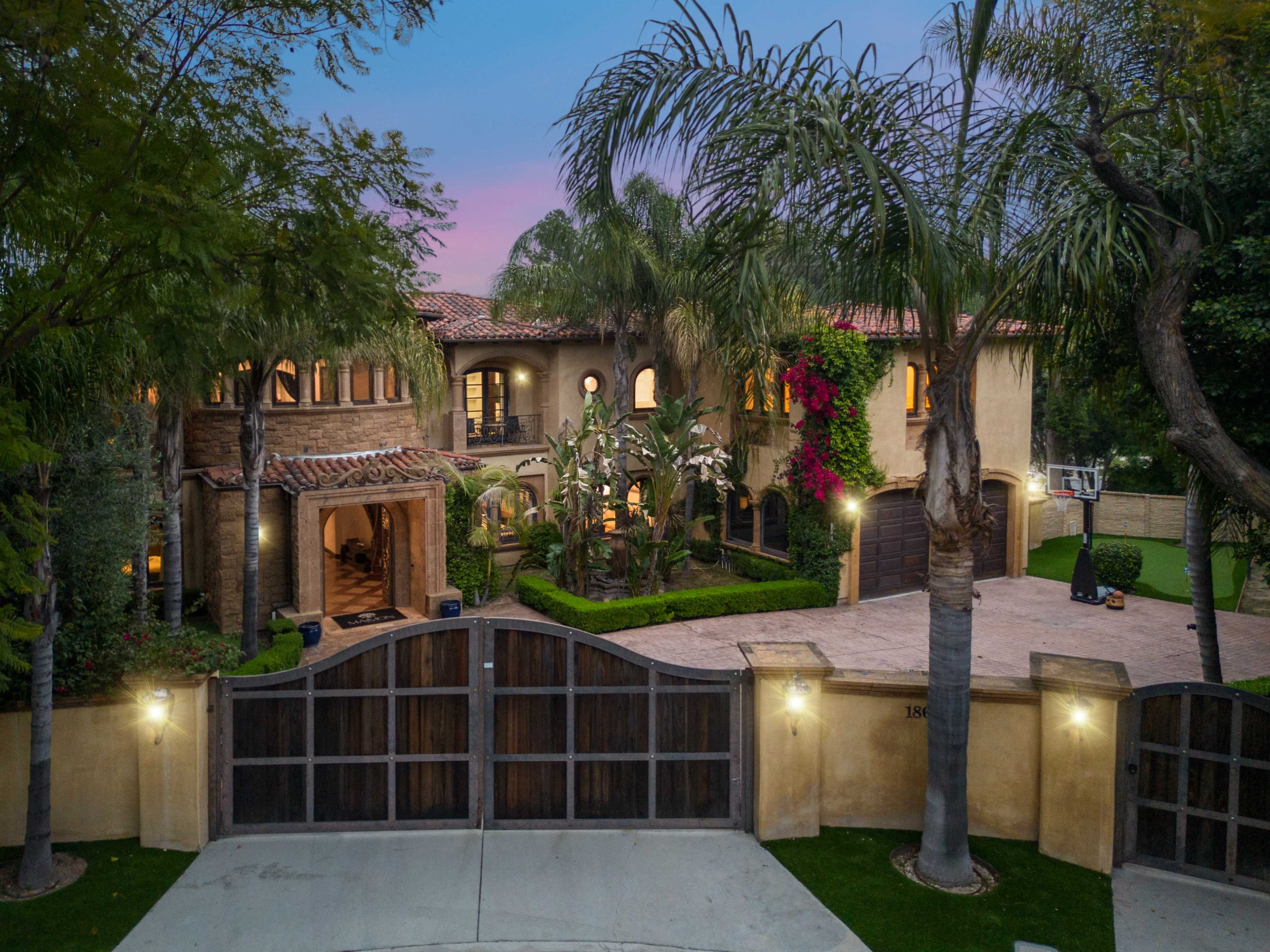 A large, two-story house with a tile roof and landscaped grounds is surrounded by palm trees and colorful flowers, featuring a driveway and a basketball hoop.