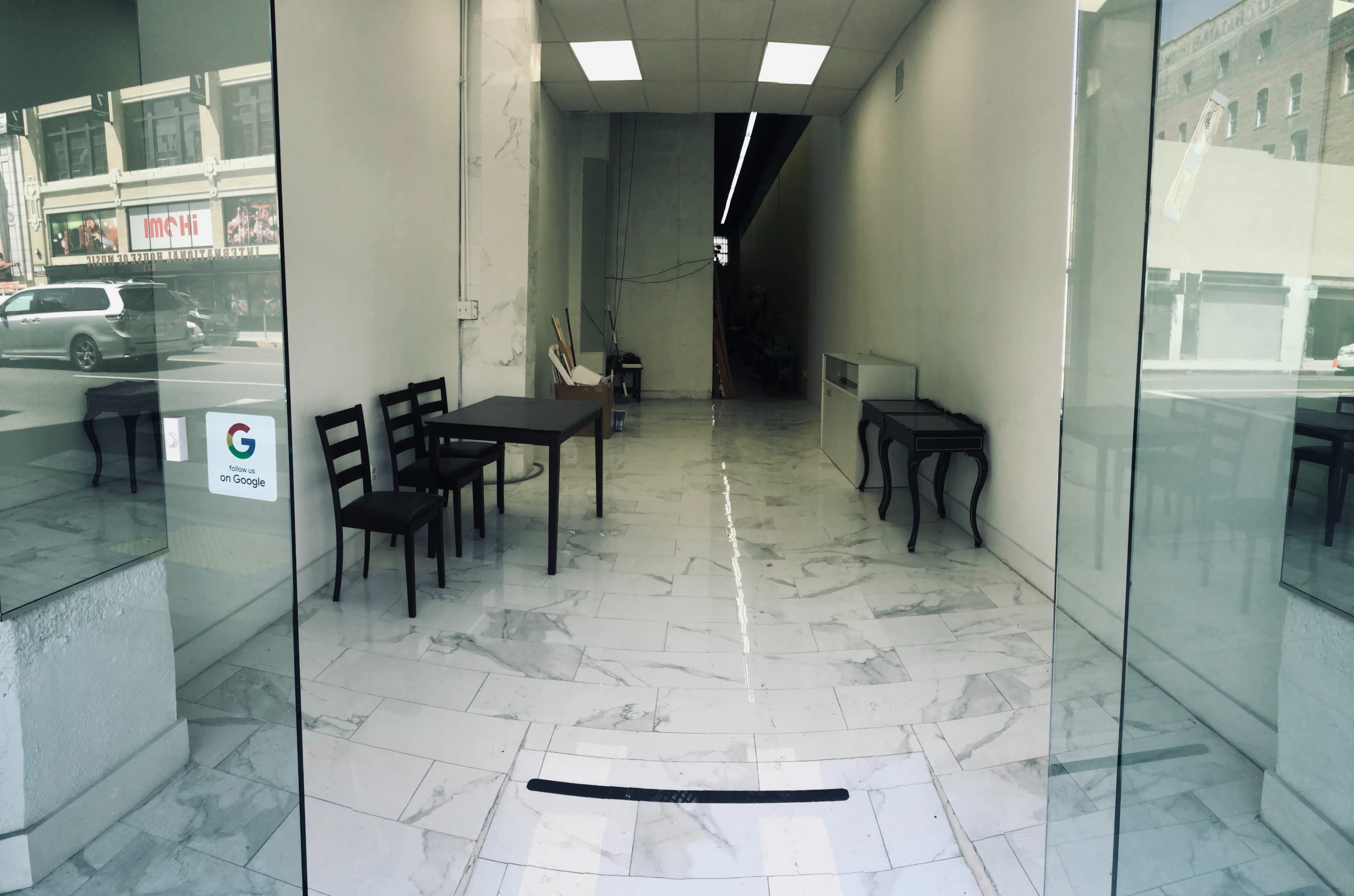 The image shows a spacious, empty room with marble flooring, featuring two chairs and a table near the entrance, and a darker corridor leading further inside.