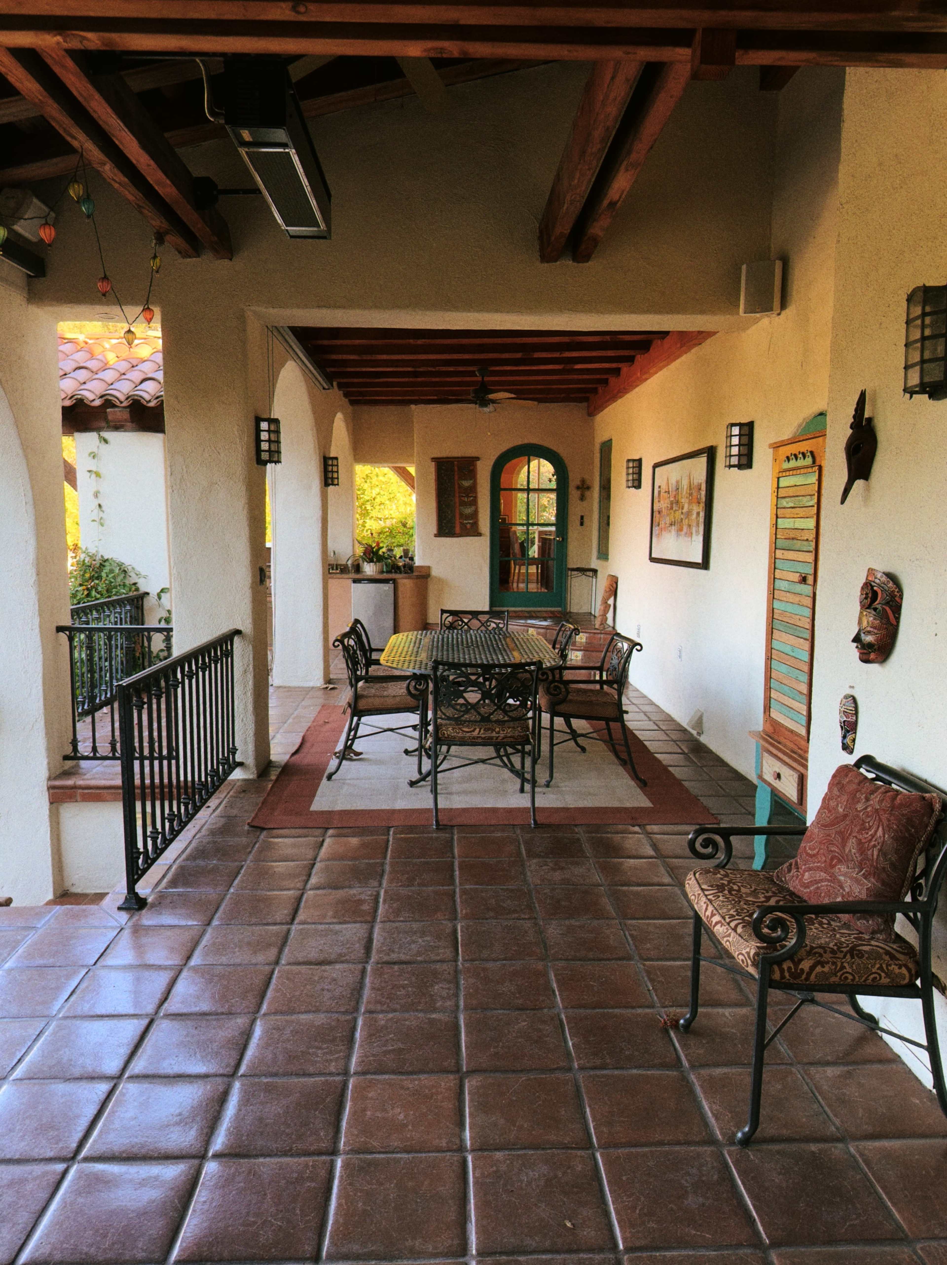 The image shows a covered porch featuring a table with chairs, decorative wall art, and tile flooring under wooden beams.